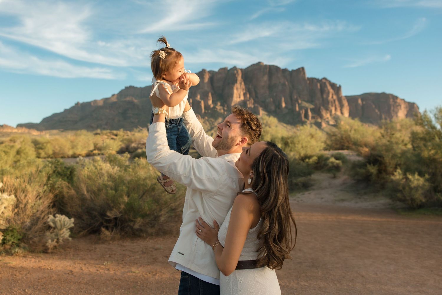 Couple holds baby up in the air in front of Superstition Mountains in Arizona desert landscape.