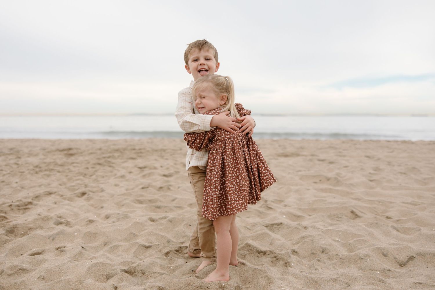 Two young children hugging and smiling on a sandy beach with ocean in background.