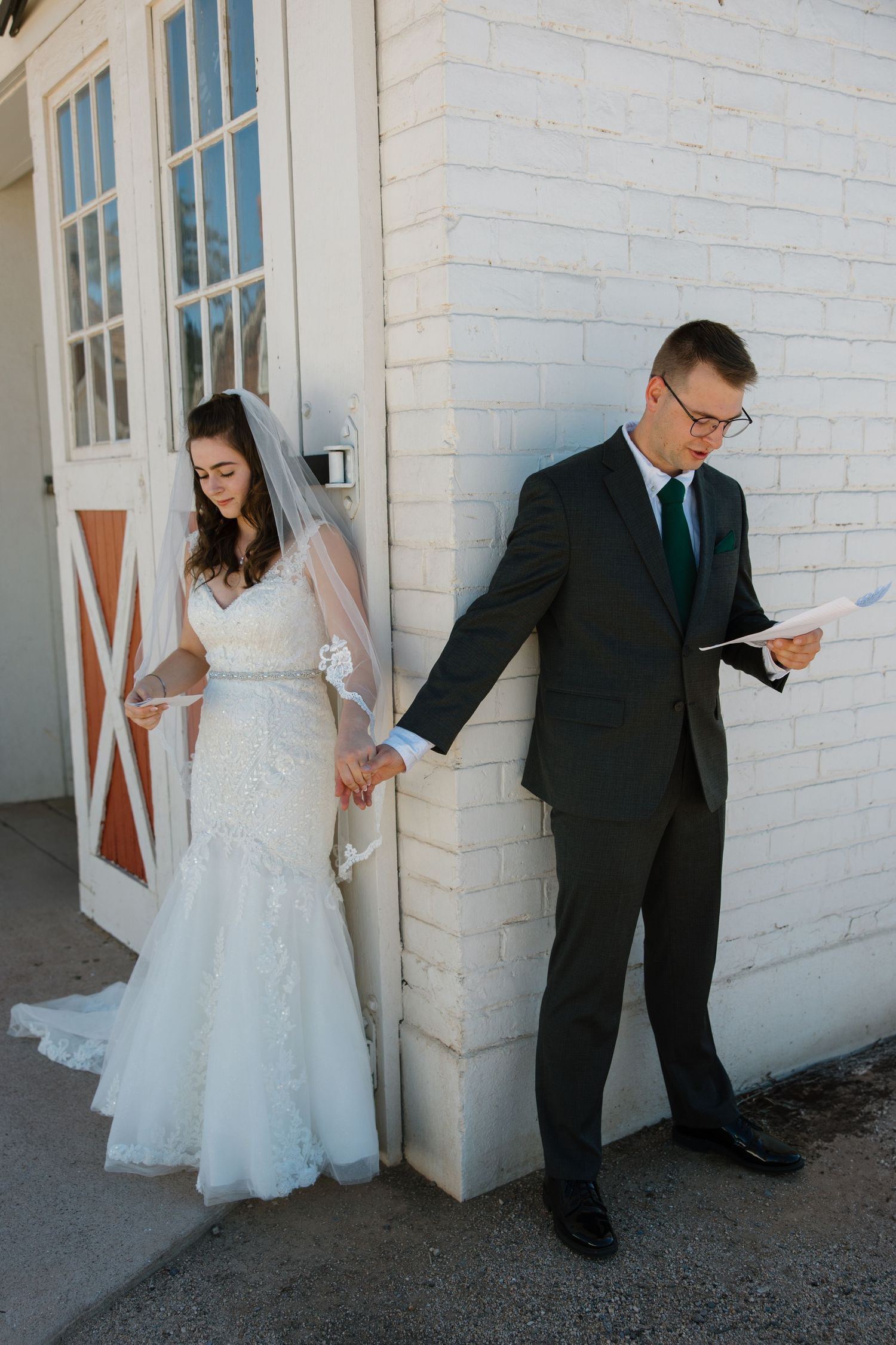 Bride and groom share private vows through a wall, holding hands outside a white brick wedding venue.