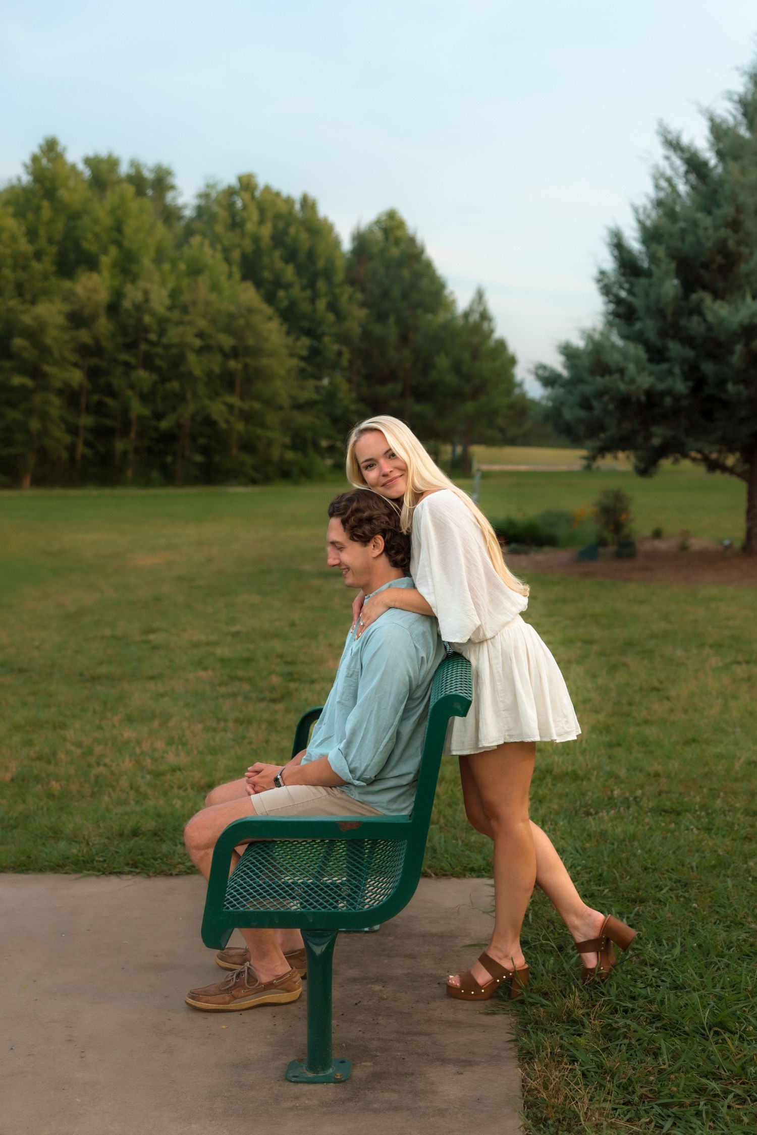 Smiling blonde woman in white skirt stands behind seated man on green park bench outdoors.
