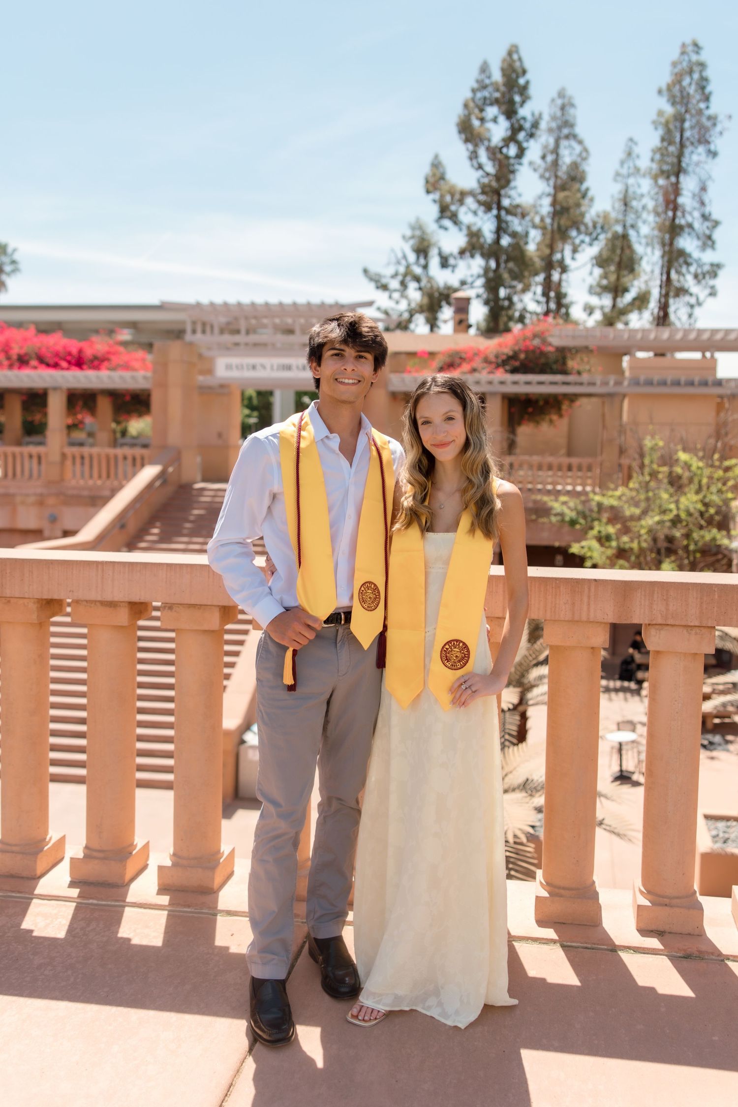 Two college graduates wearing gold stoles pose together on a sunny campus terrace.
