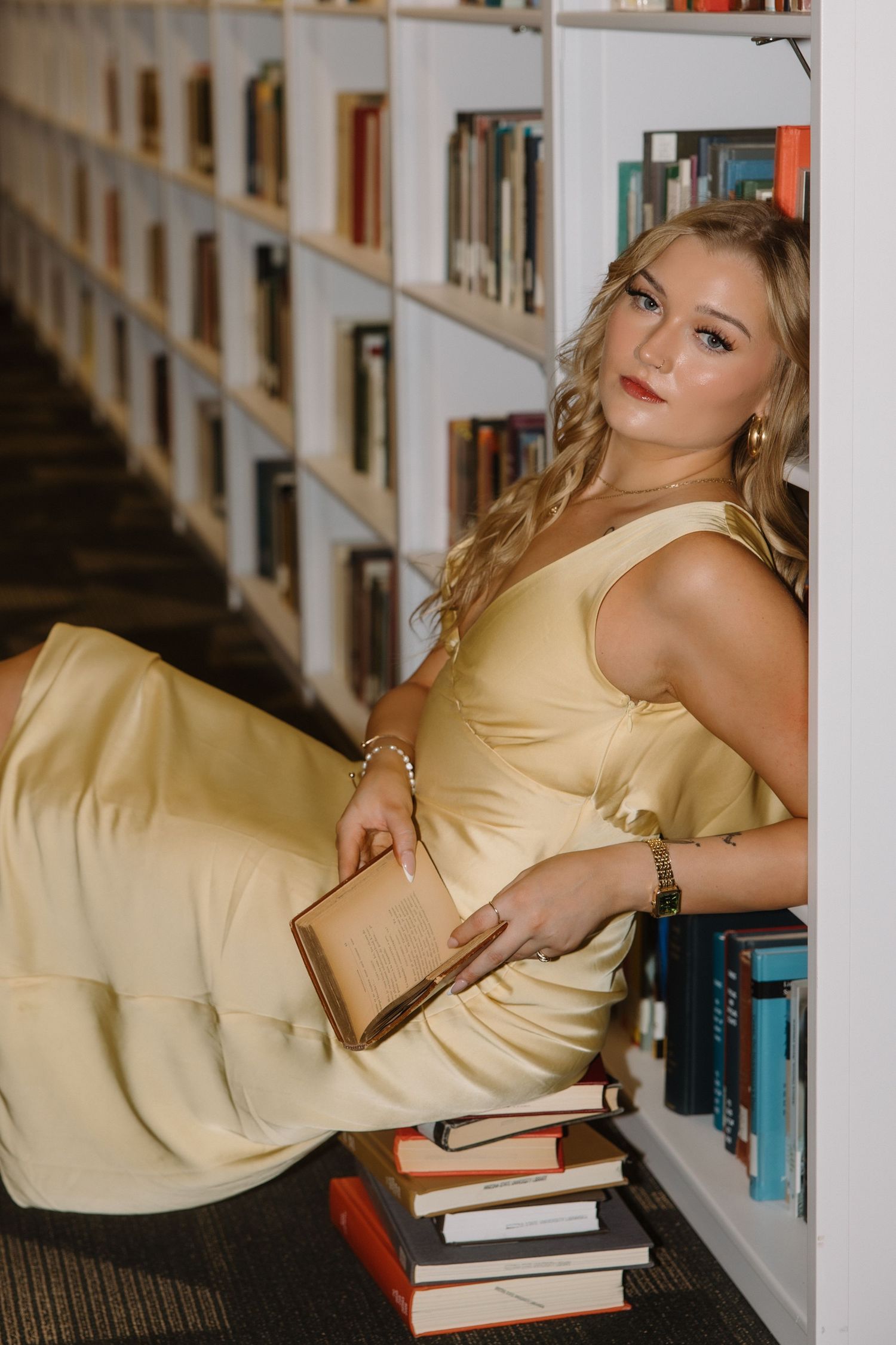 Young blonde woman in yellow satin dress poses with books in a library.