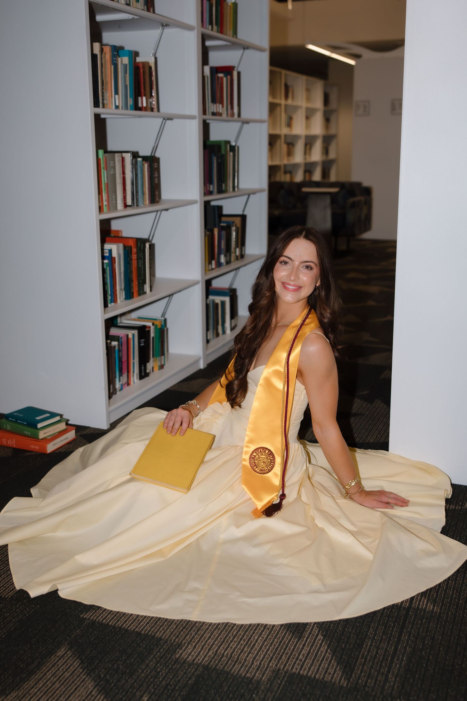A smiling woman in a yellow graduation gown and stole sits on a library floor holding a book.