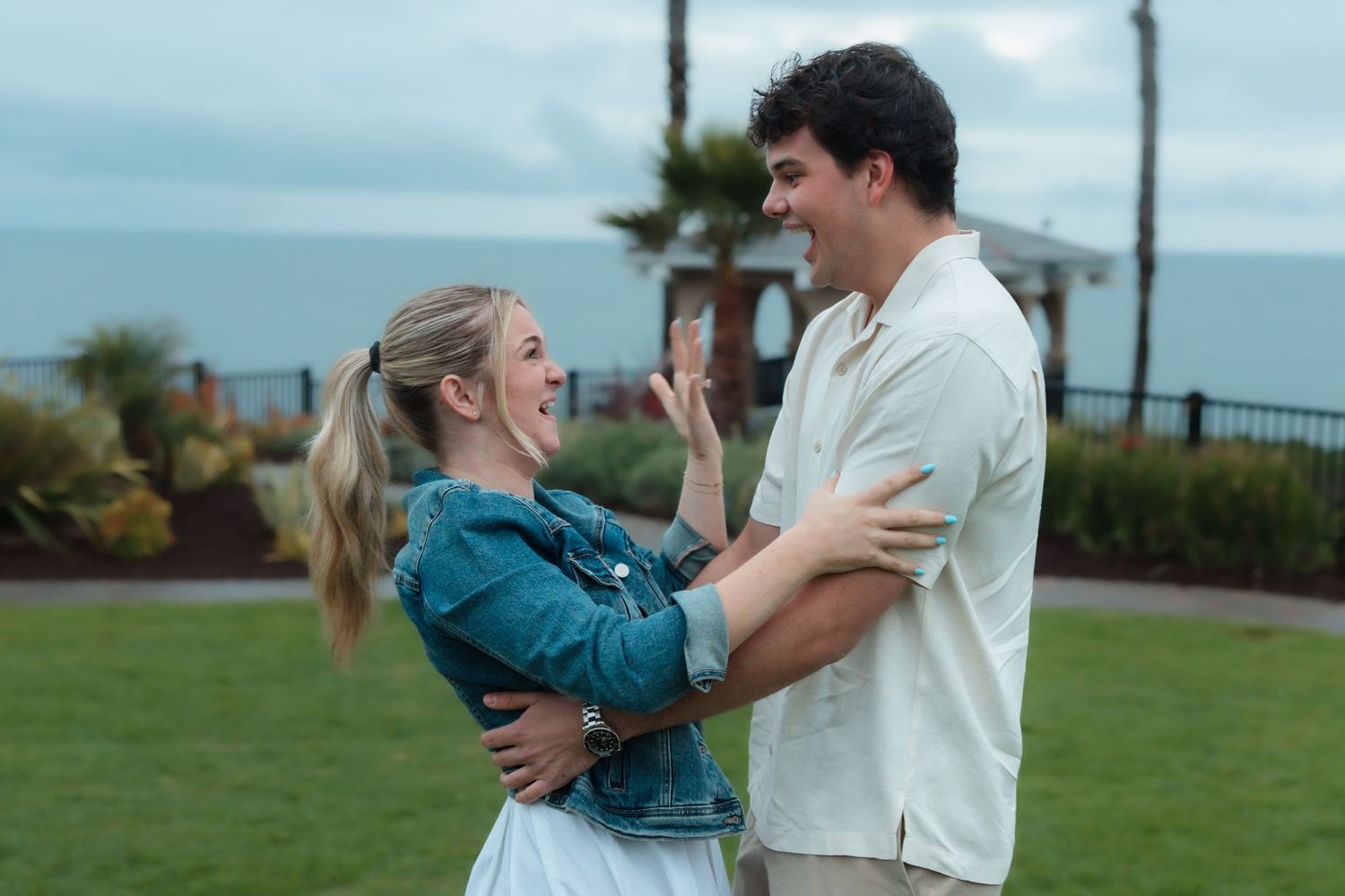 A joyful couple laughing and embracing outdoors near a coastal park with palm trees and cloudy sky.