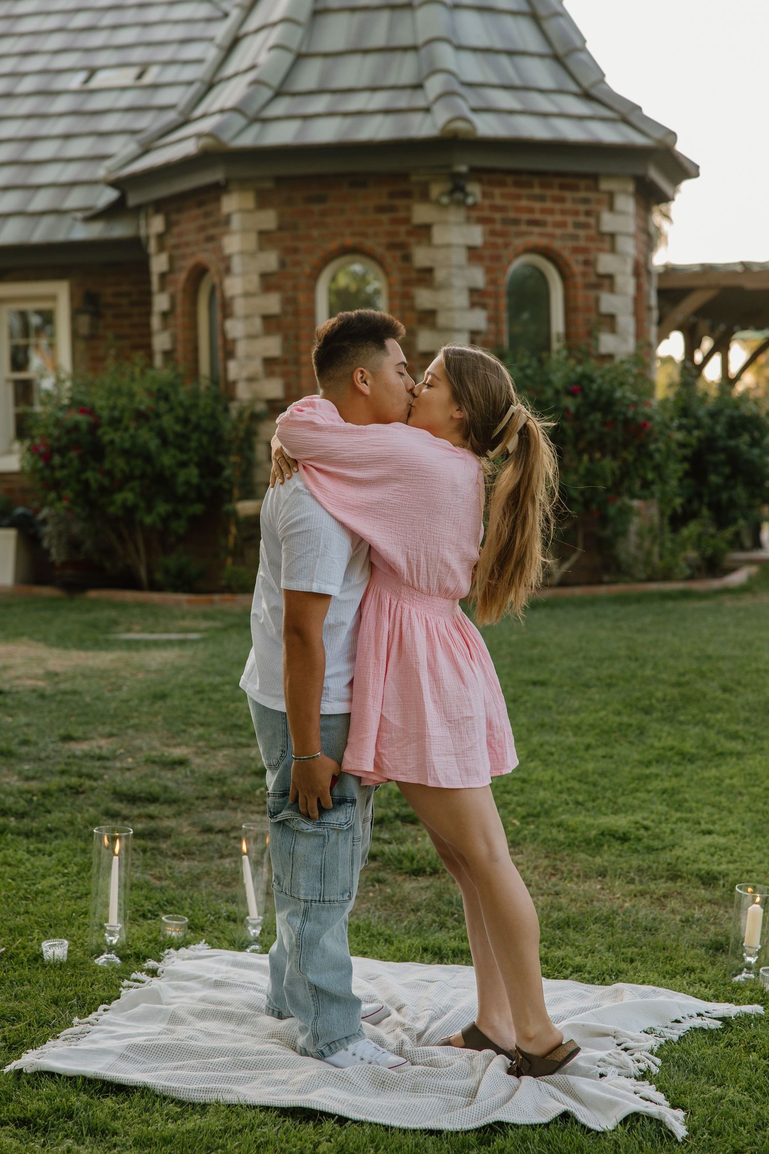Couple sharing a romantic kiss on a white blanket with candles in a garden setting near a charming brick building.