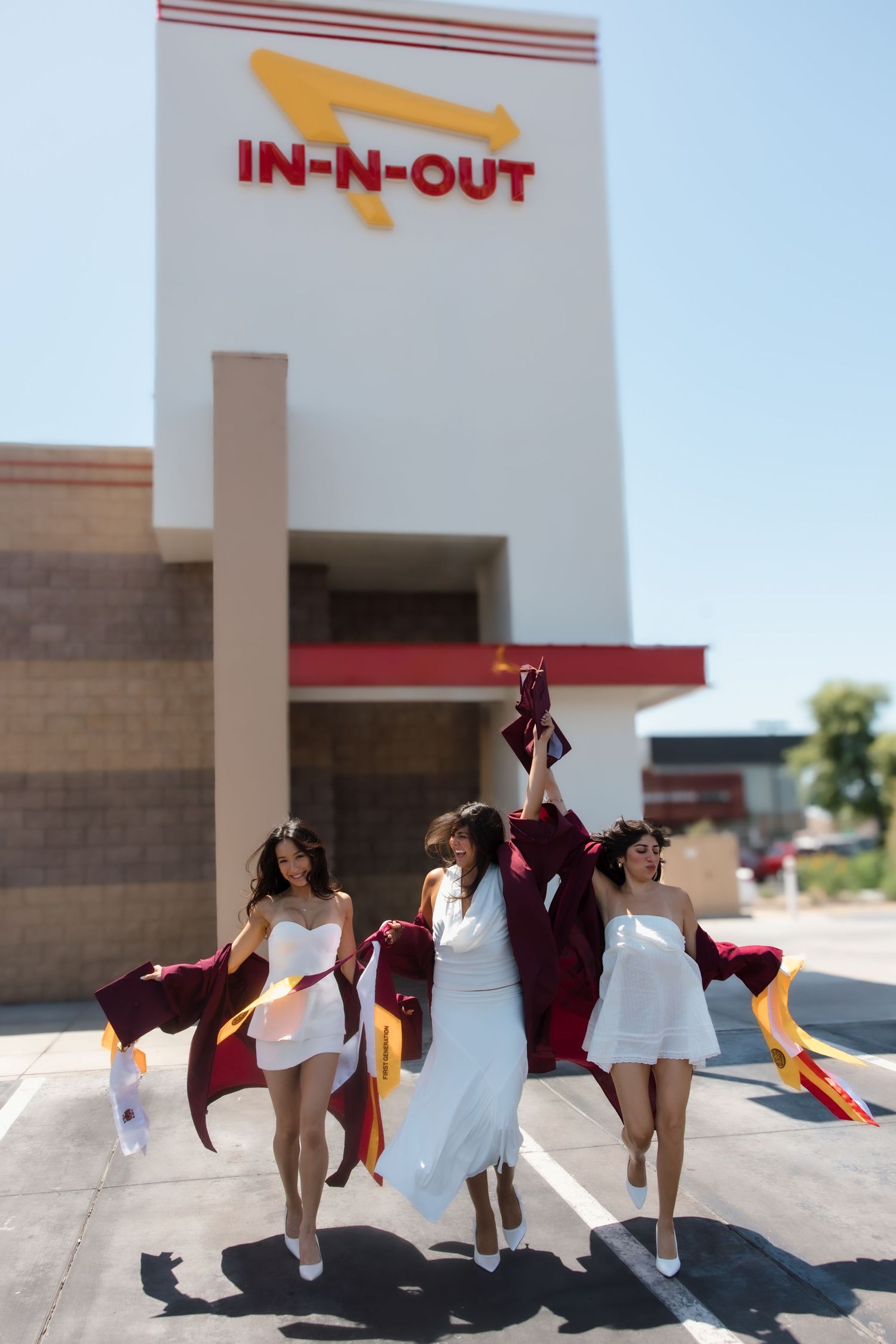 Three graduates in white dresses celebrating with raised arms outside In-N-Out Burger.