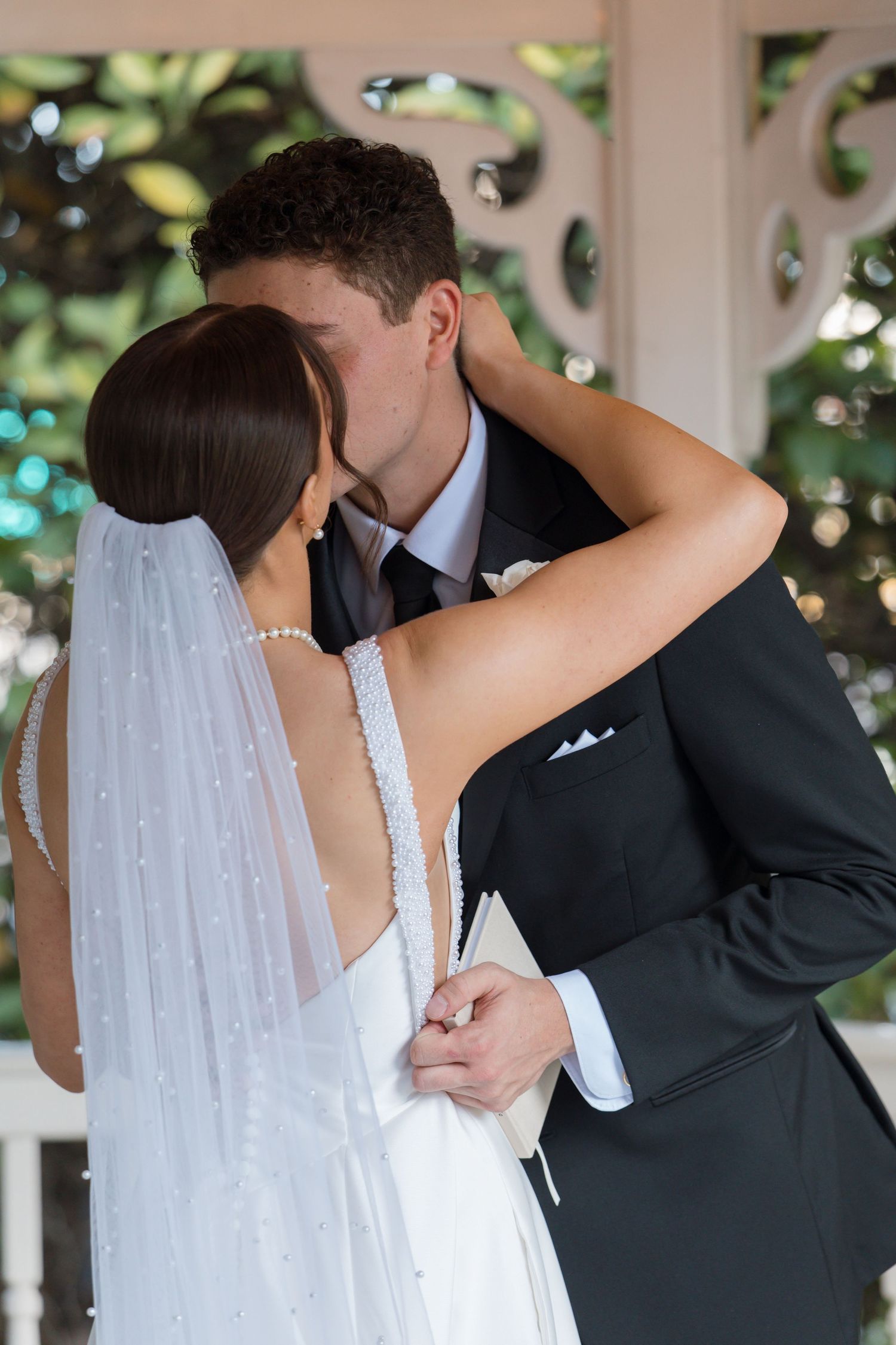 Newlyweds share first kiss embrace under white garden gazebo surrounded by lush greenery.