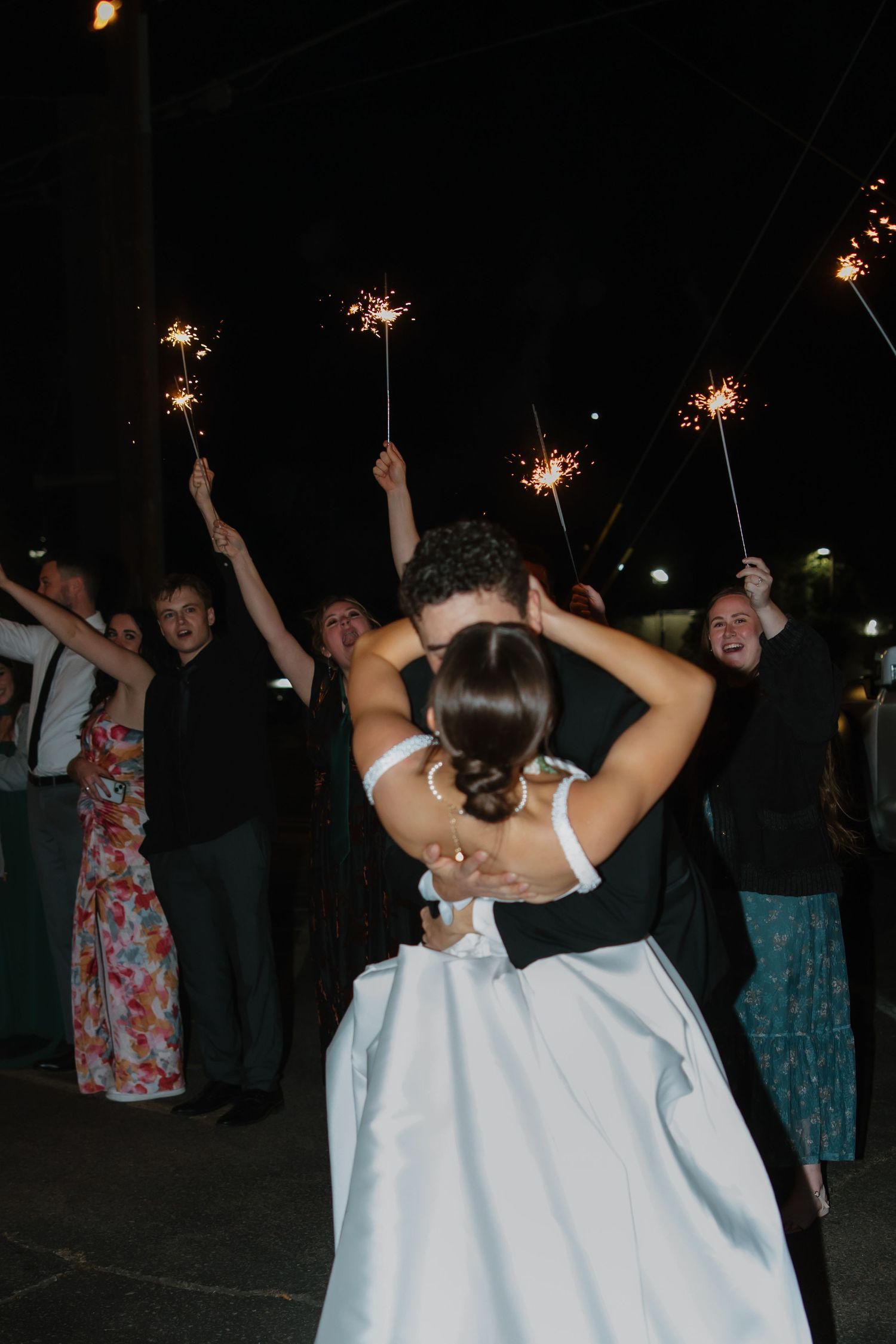 Newlyweds share a kiss surrounded by guests holding sparklers during festive outdoor wedding sendoff at night.