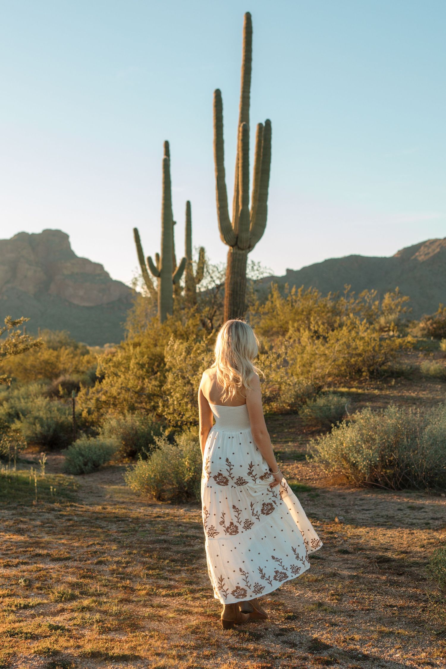 Back view of blonde woman in white floral maxi dress walking through Sonoran desert at golden hour.