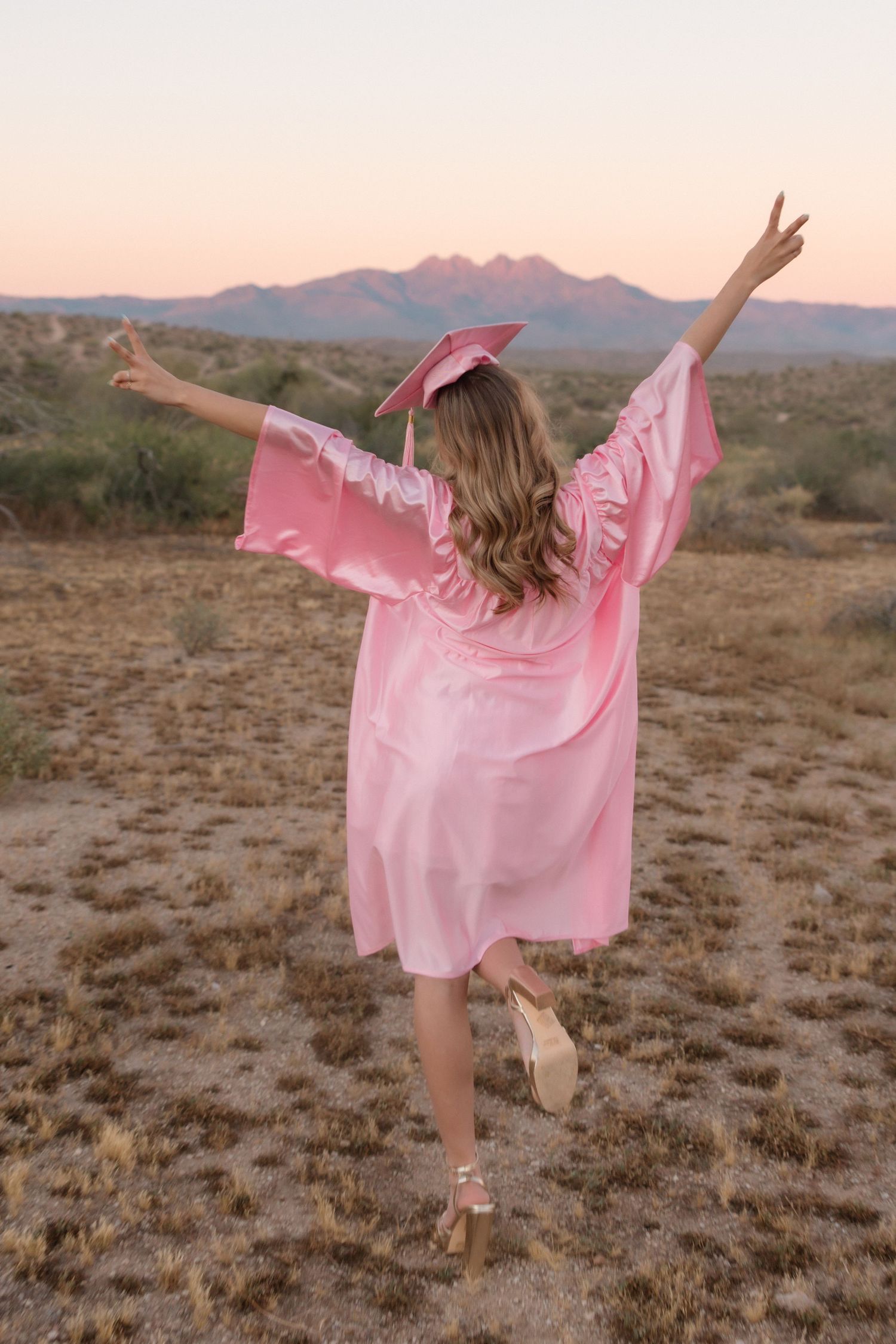 Graduate in pink cap and gown celebrating with arms raised in desert landscape at golden sunset.