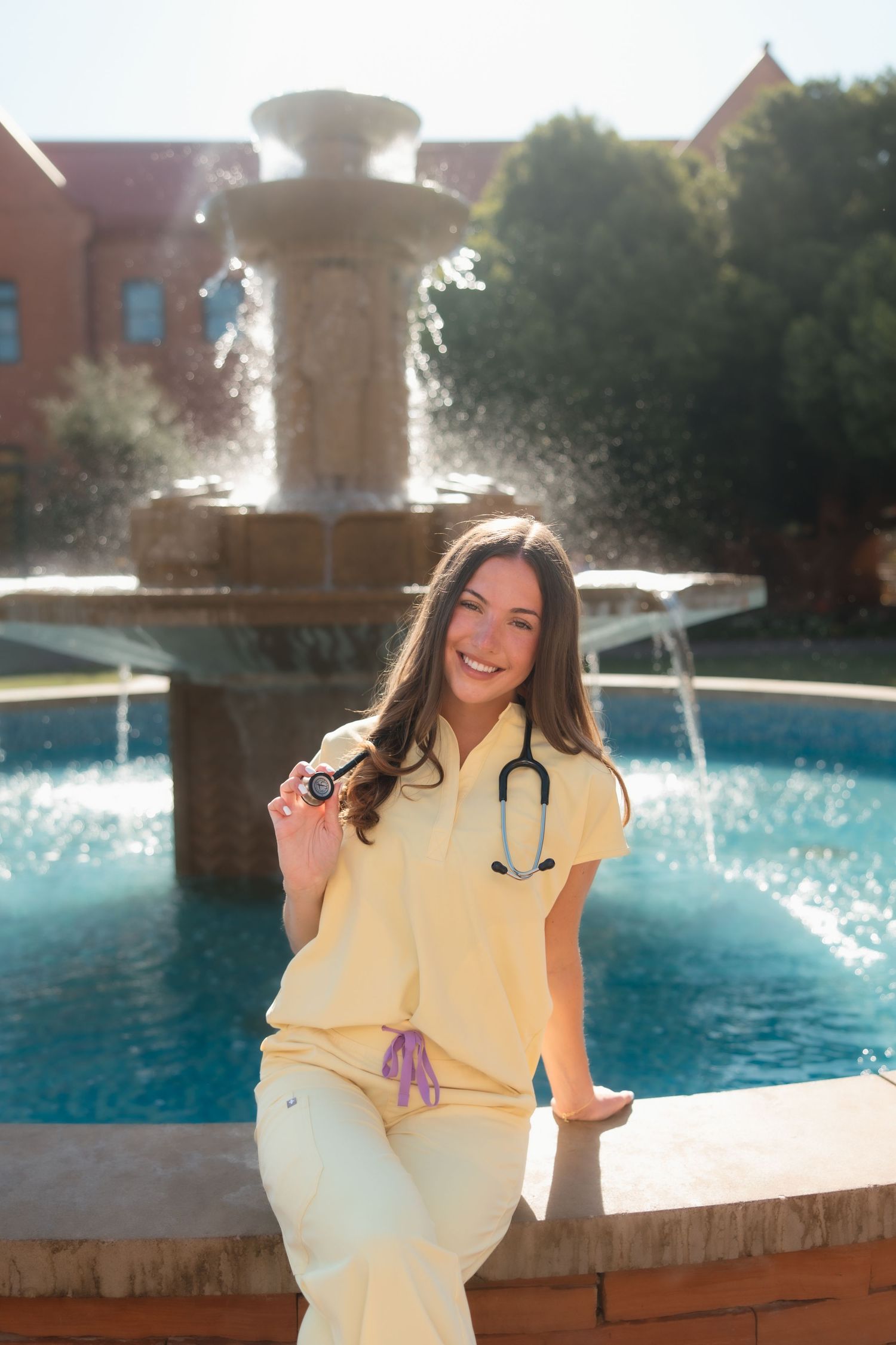 Happy nursing graduate in yellow scrubs with stethoscope sitting by ornate campus fountain.