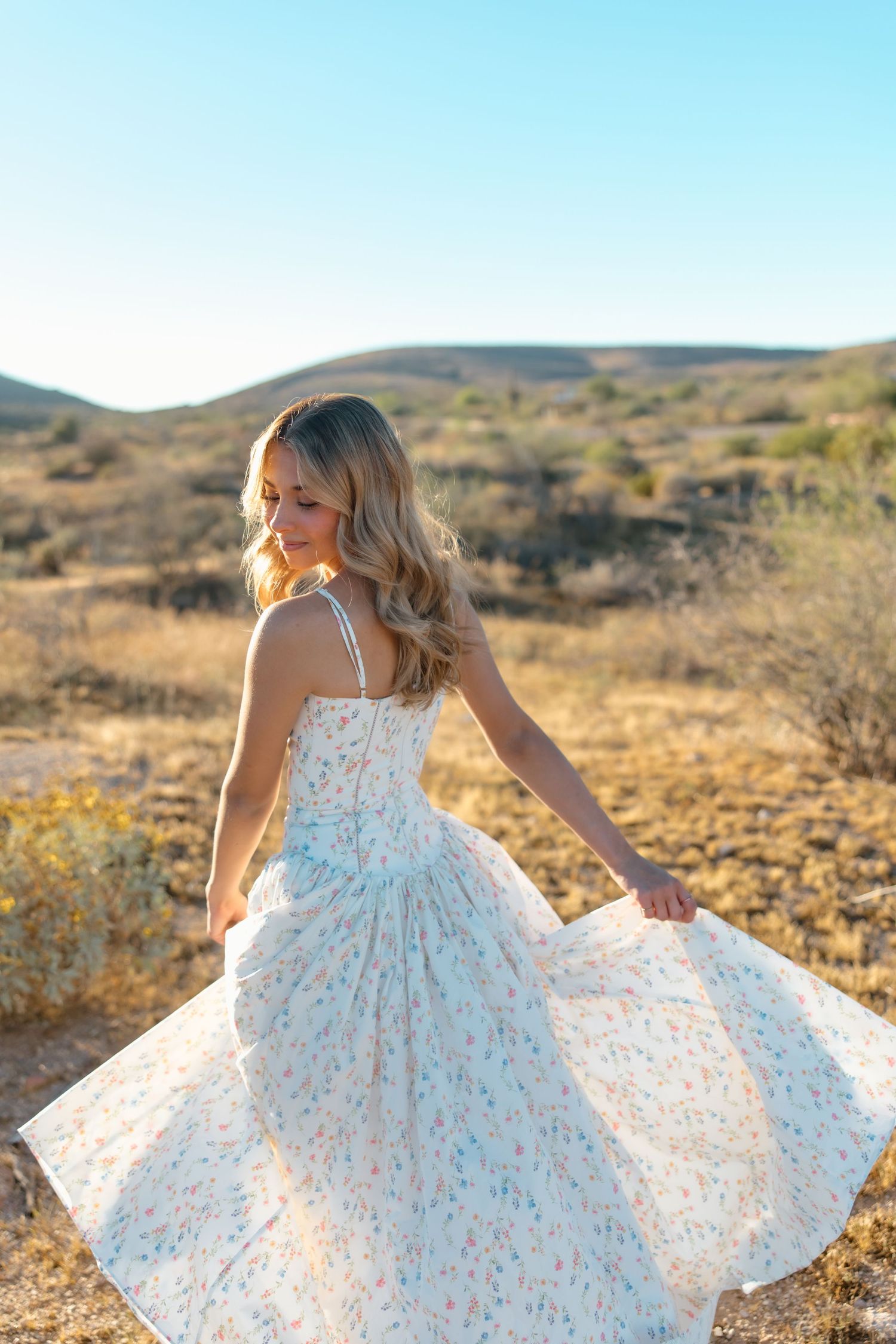 Blonde woman twirling in a white floral sundress in a sunny desert landscape with rolling hills.