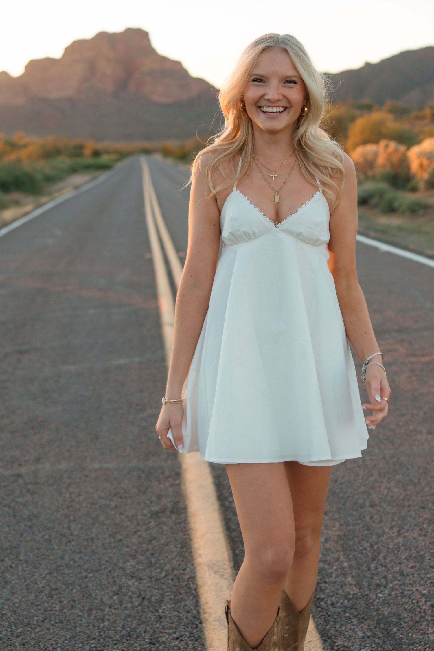 Smiling blonde woman in white mini dress and cowboy boots standing on open road with mountain backdrop at sunset.