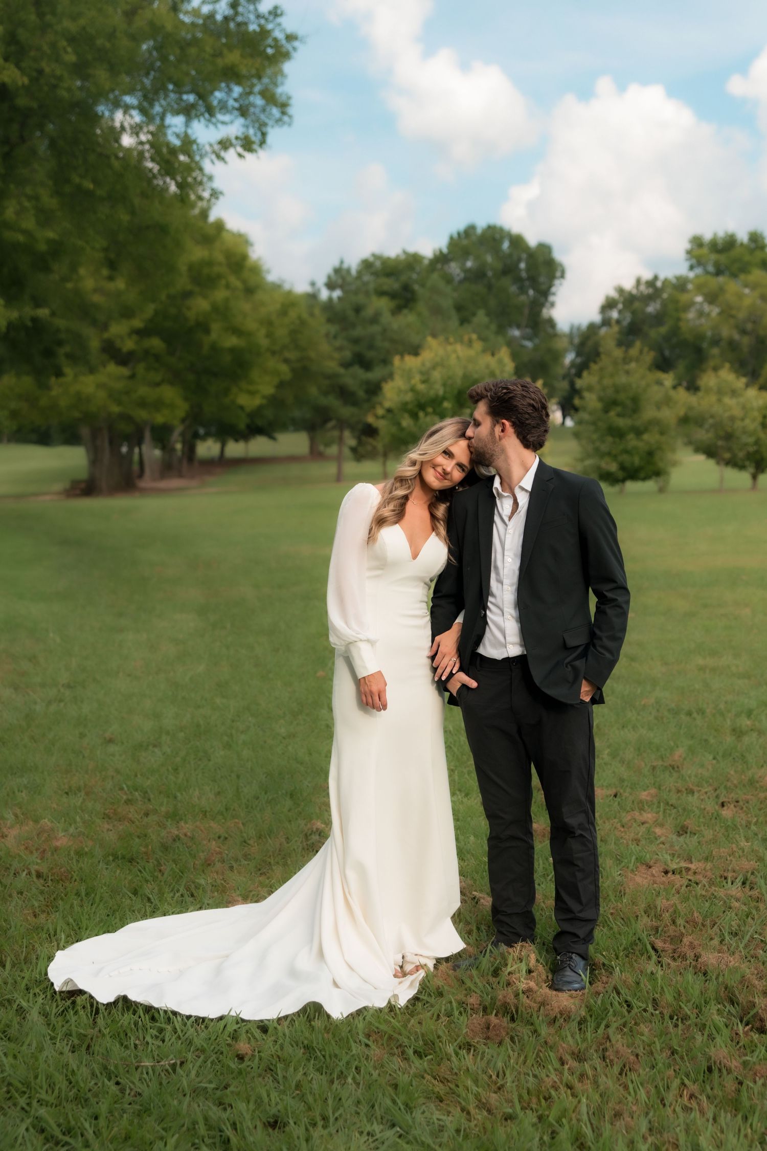 Bride in white gown and groom in black suit share a tender kiss in a lush green outdoor setting.