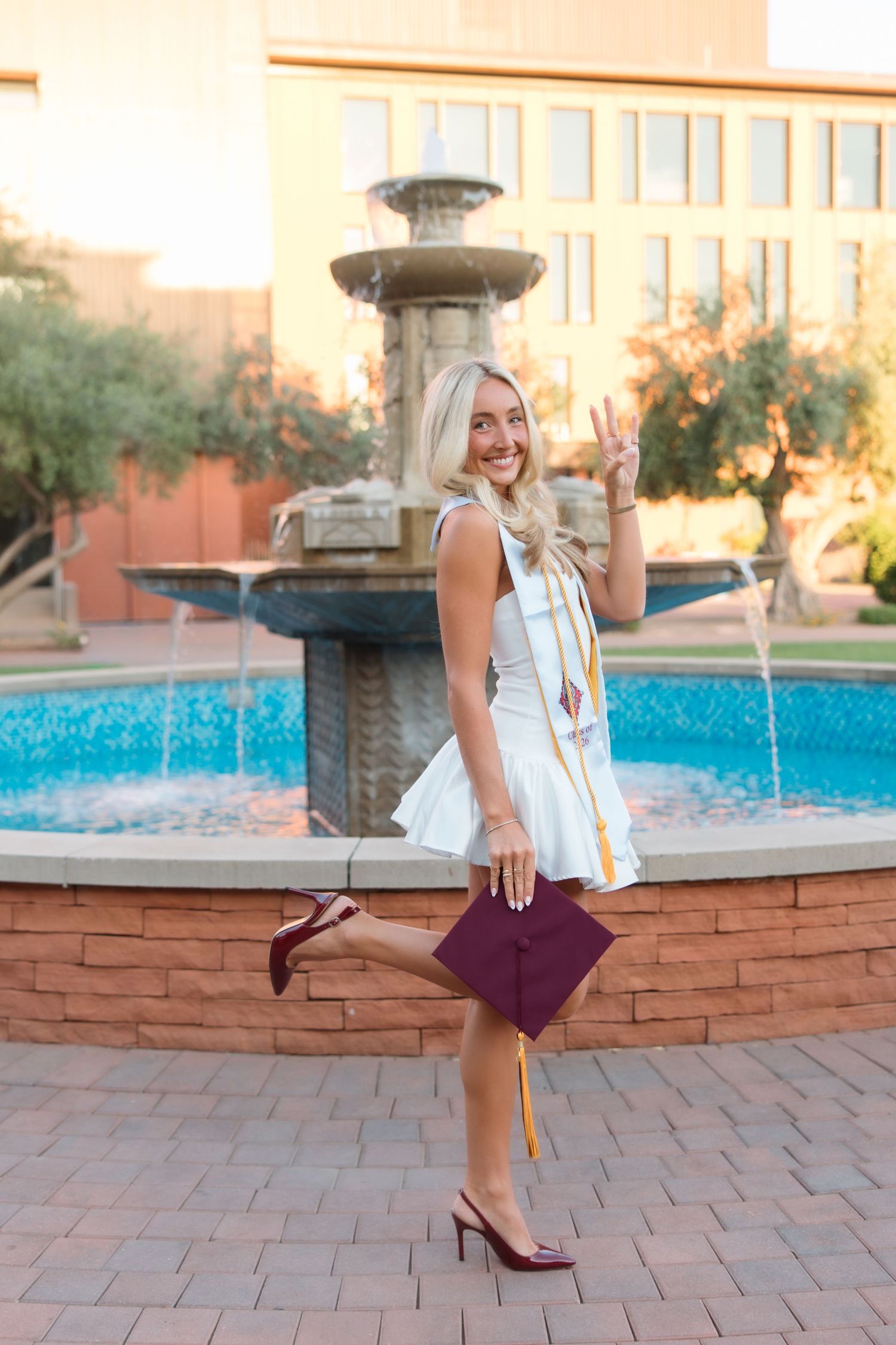 Blonde female graduate in white dress holds maroon cap by fountain, flashing peace sign in golden hour light.