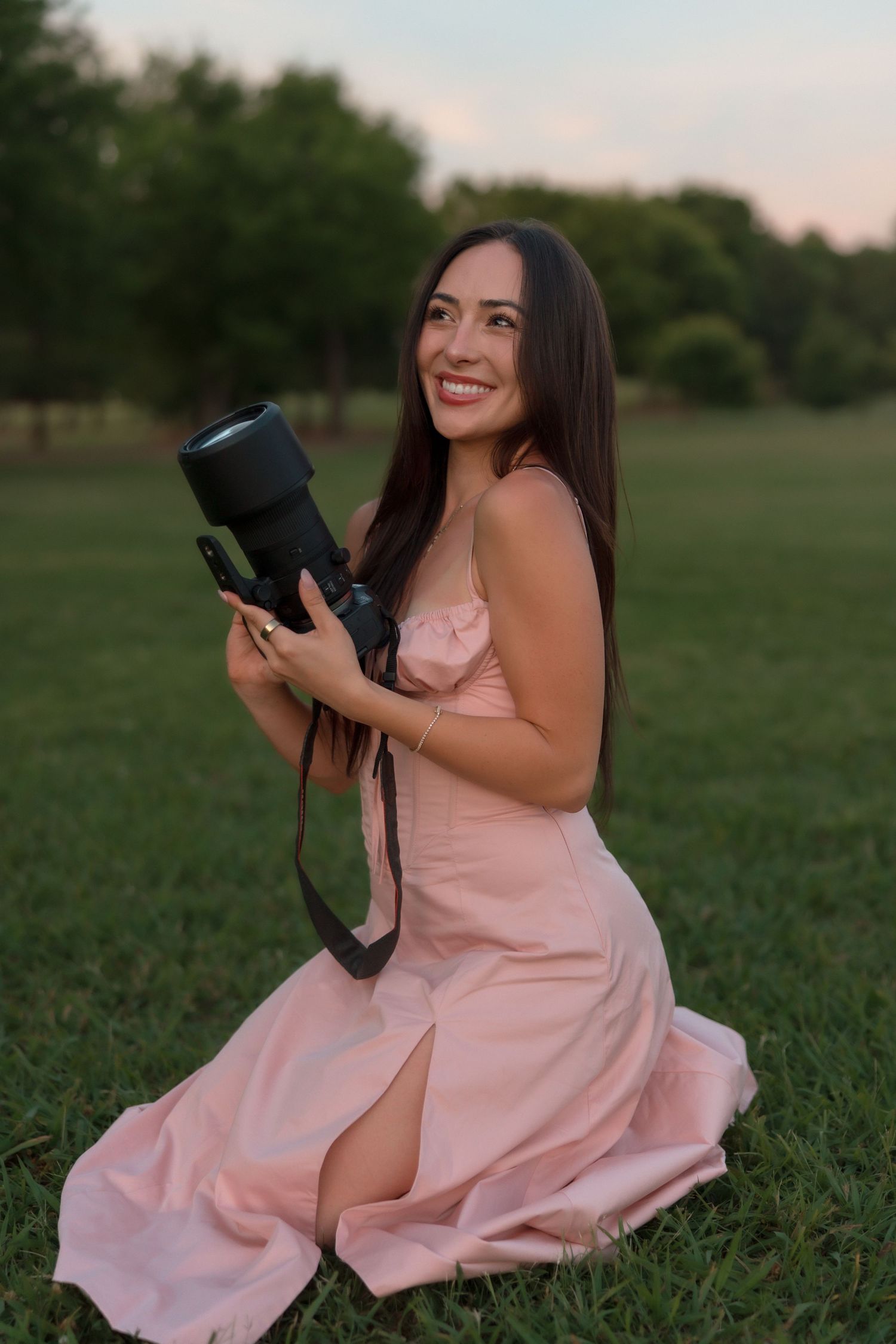 Smiling brunette photographer in pink dress kneeling on green grass holding professional camera.