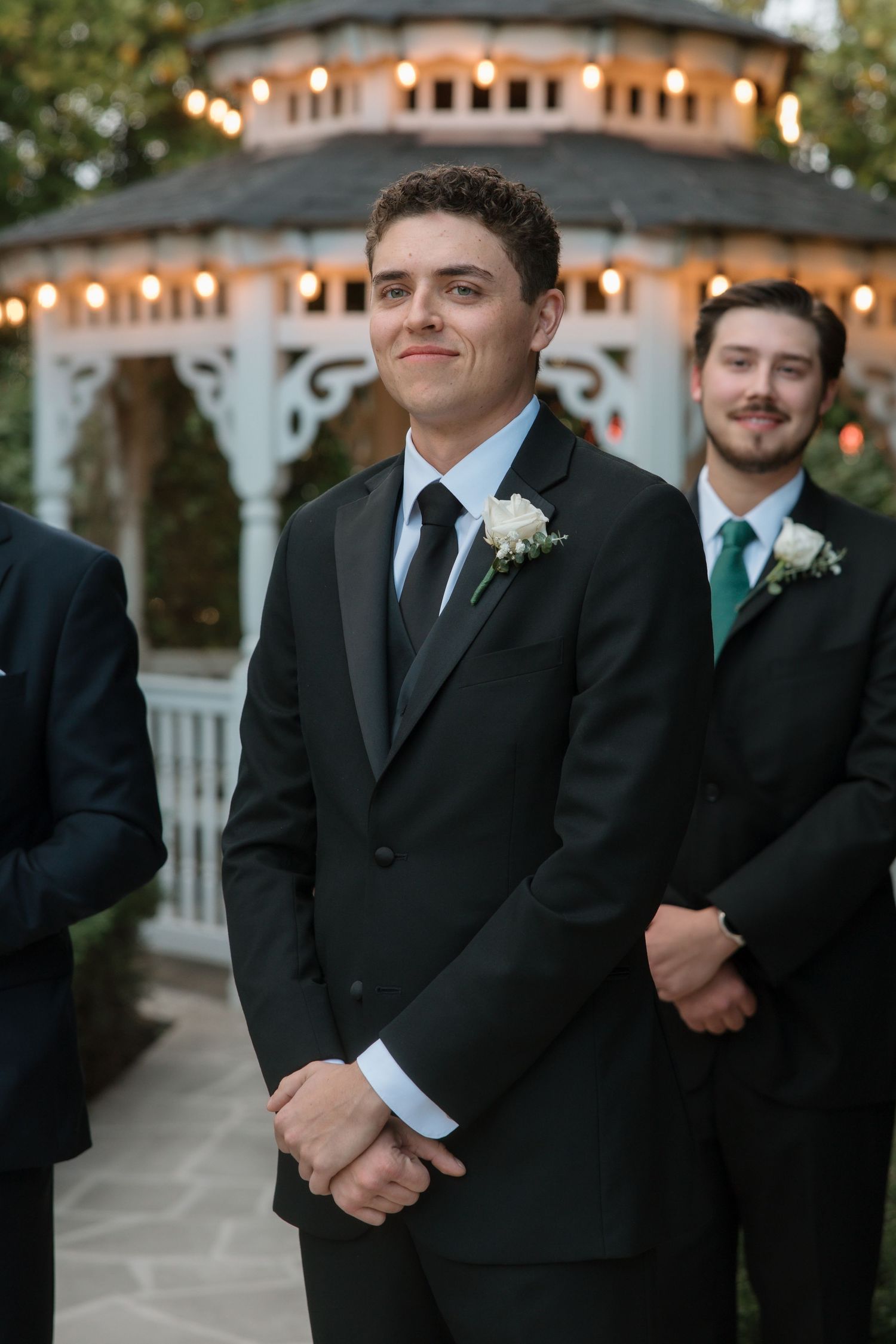 Groom in black tuxedo with white boutonniere smiling at wedding ceremony near lit gazebo.