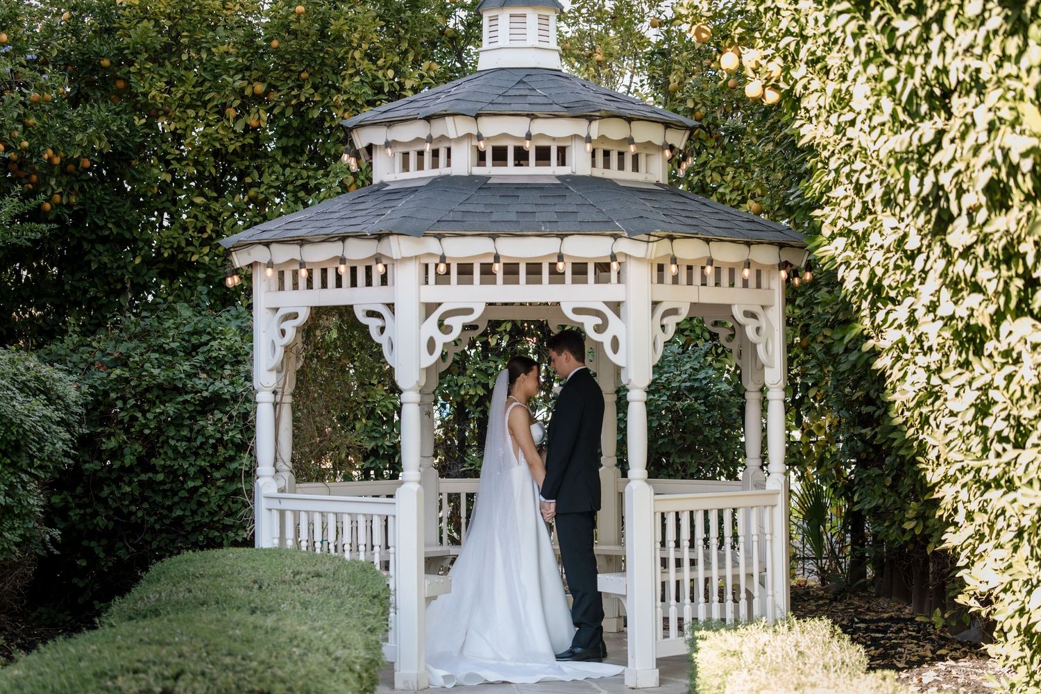 Bride and groom share a romantic moment under a white octagonal gazebo surrounded by autumn foliage.