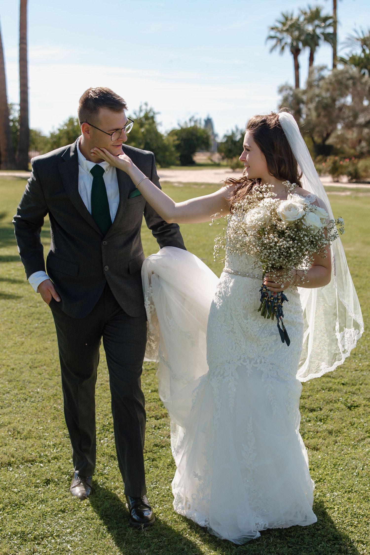 Bride touches grooms chin affectionately as they walk together on green grass holding a white bouquet.