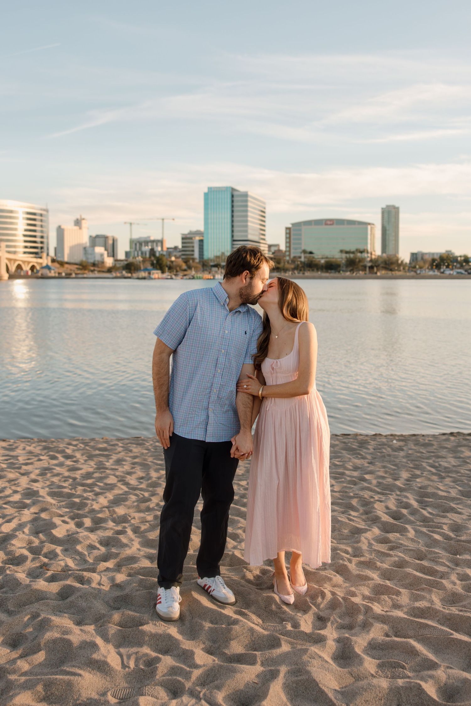 Couple sharing a kiss on a sandy beach with a city skyline and calm water in the background at golden hour.