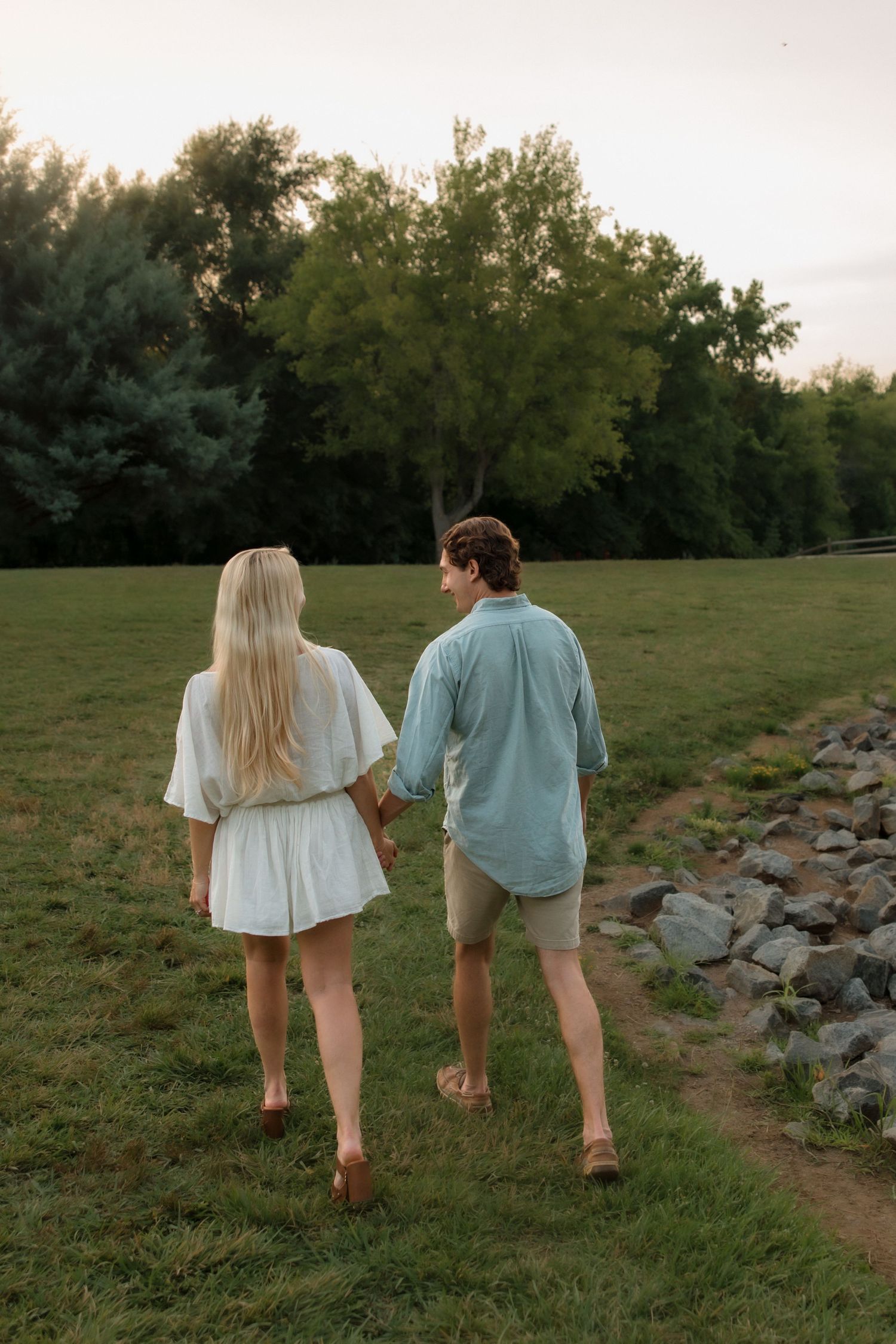 Couple holding hands walking barefoot on green grass toward trees at sunset near a rocky shoreline.