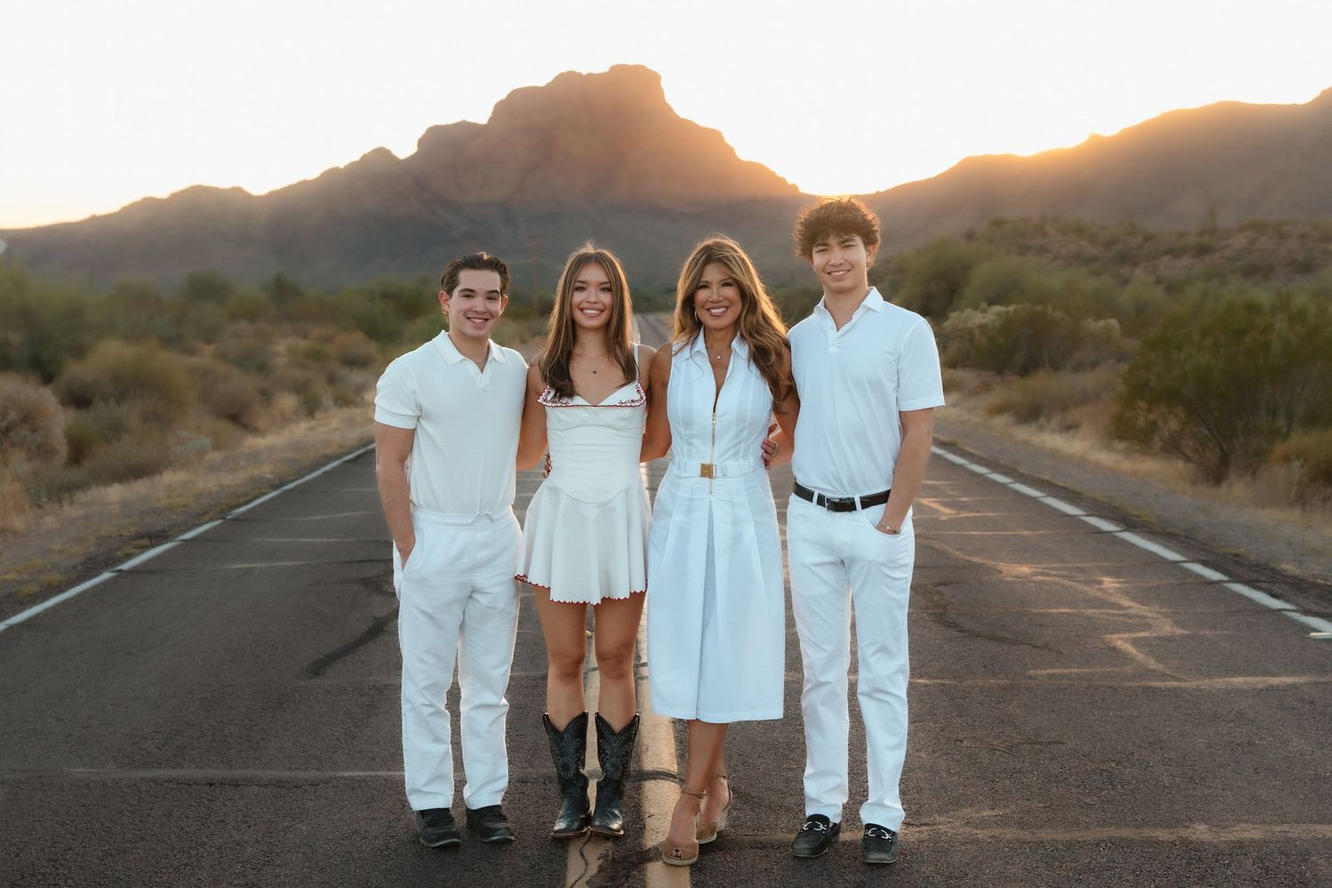 Family of four posing in white outfits on desert road with mountain backdrop at sunset.