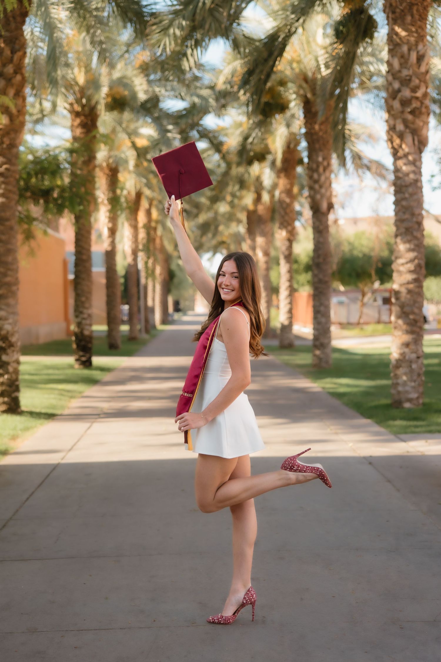 Joyful graduate in white dress raises maroon cap celebrating along sunny palm tree-lined campus path.