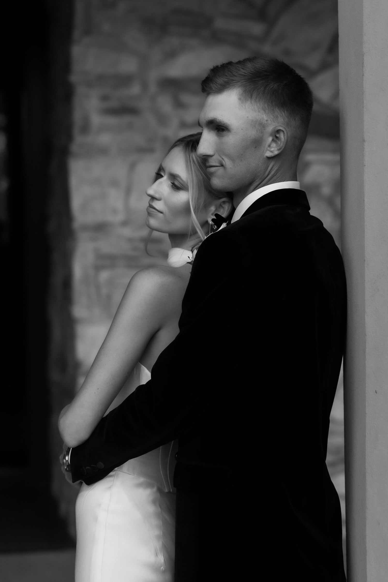 Elegant black and white portrait of bride and groom embracing against stone wall at wedding venue.