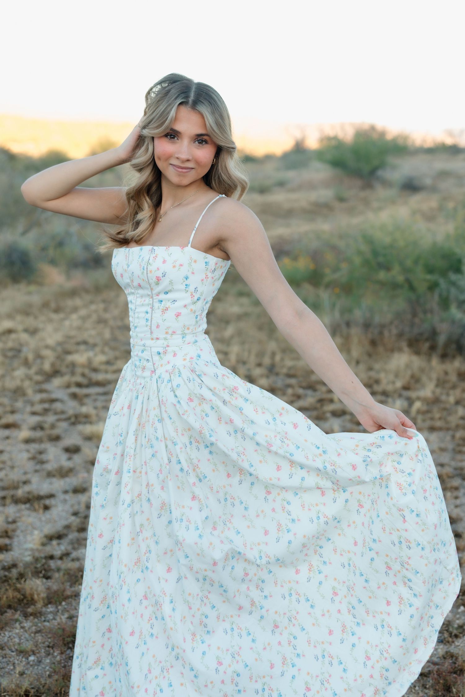 Blonde woman in white floral maxi dress posing gracefully in desert at golden hour.