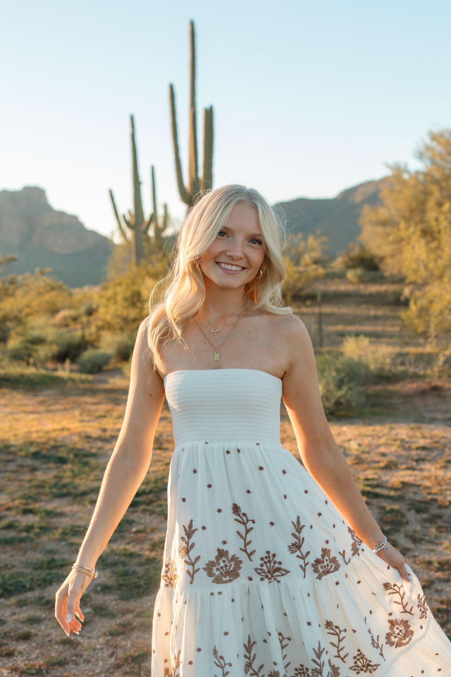 Smiling blonde woman in white strapless embroidered dress posing in Arizona desert at golden hour.