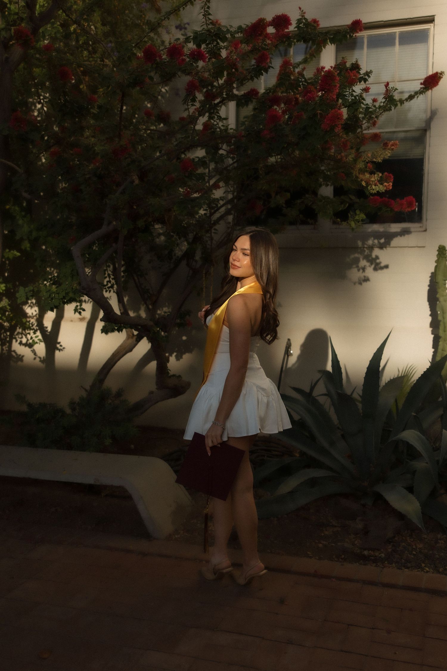 Young woman in white skirt and yellow top poses outdoors near red flowering tree and white building at dusk.