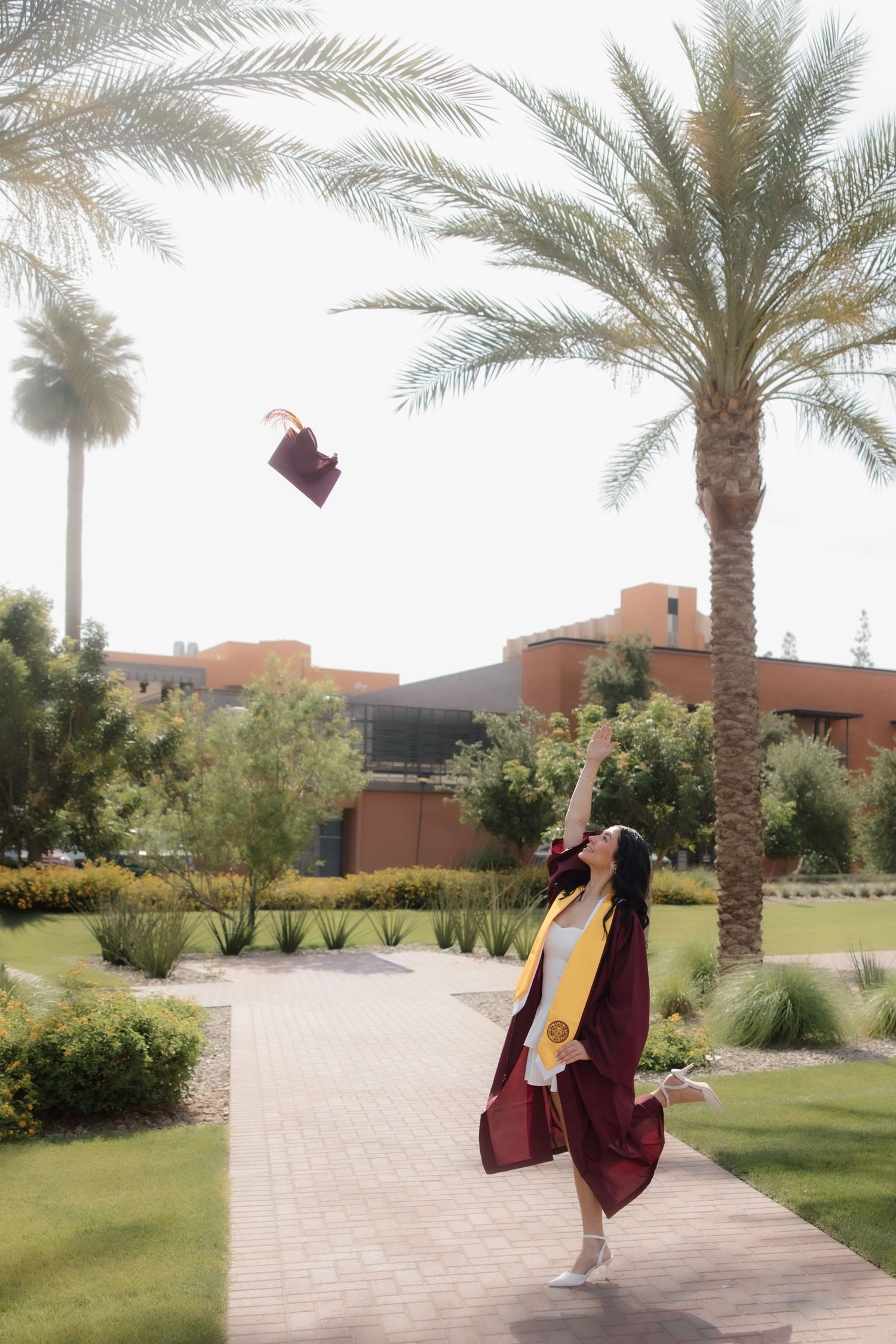Graduate in maroon cap and gown tossing mortarboard into air beside palm trees on sunny campus walkway.