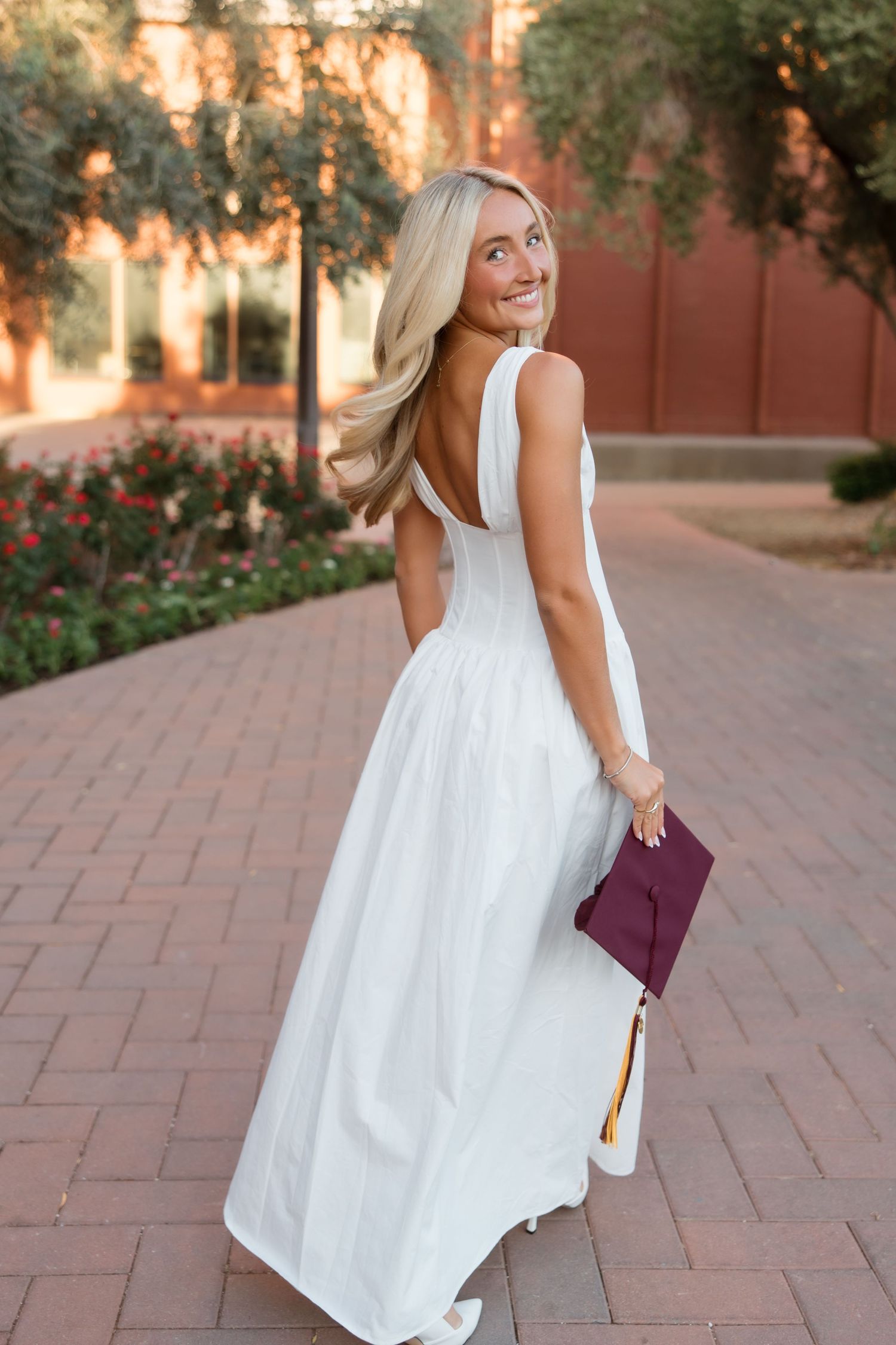 Smiling blonde woman in white backless gown holding maroon graduation cap on brick path.