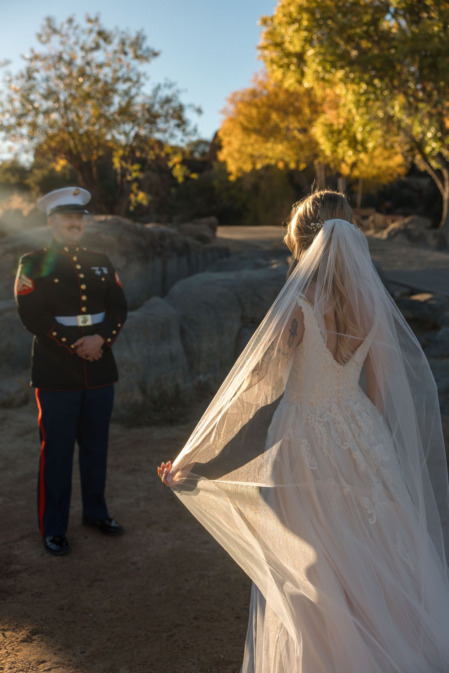 US Marine groom watches beautiful bride in lace wedding dress with flowing veil during outdoor sunset ceremony.