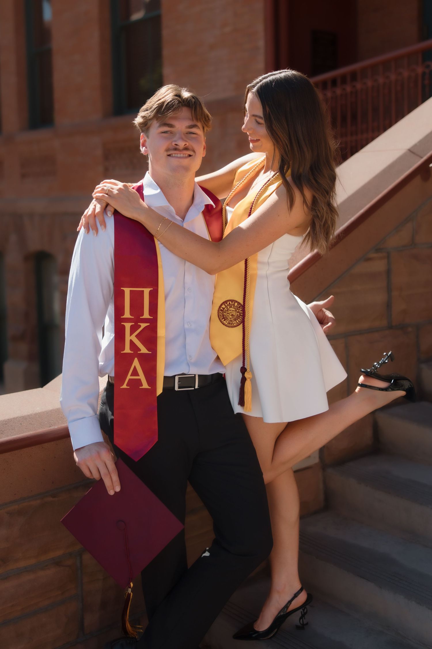 College graduates pose together on brick building steps, wearing Pi Kappa Alpha and honor stoles.