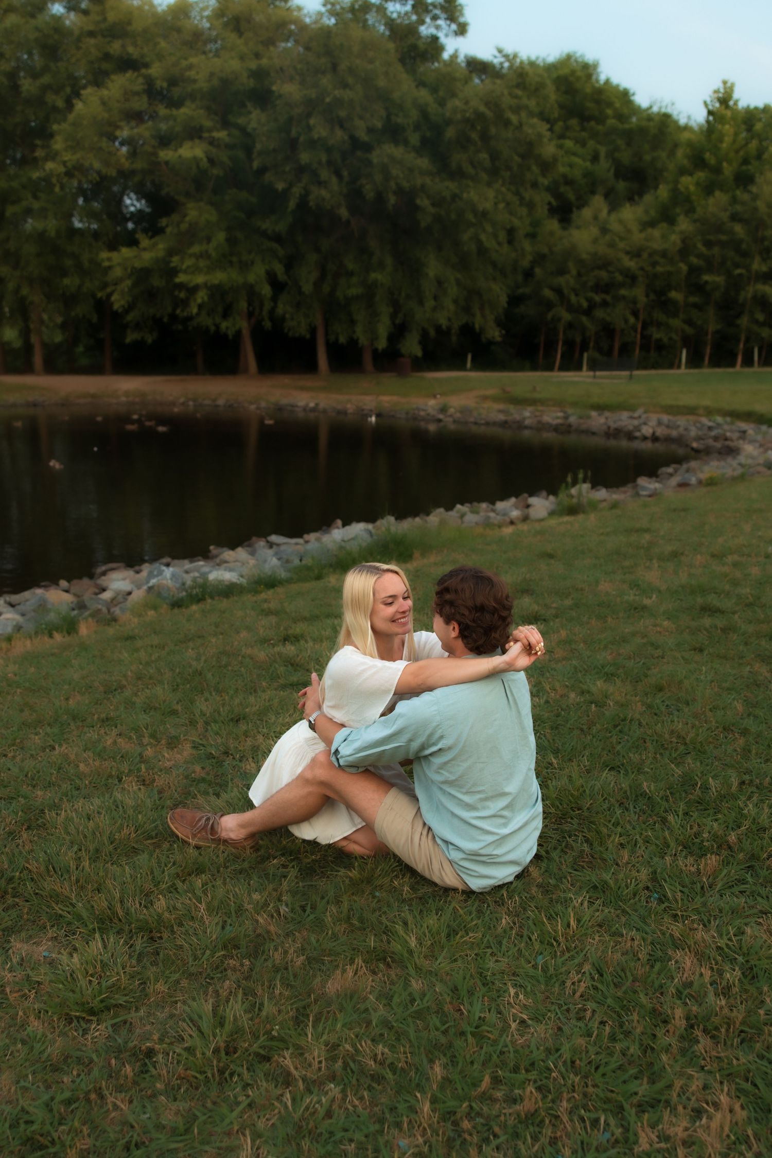 Couple sitting on grass by a lake, woman smiling while embracing her partner.