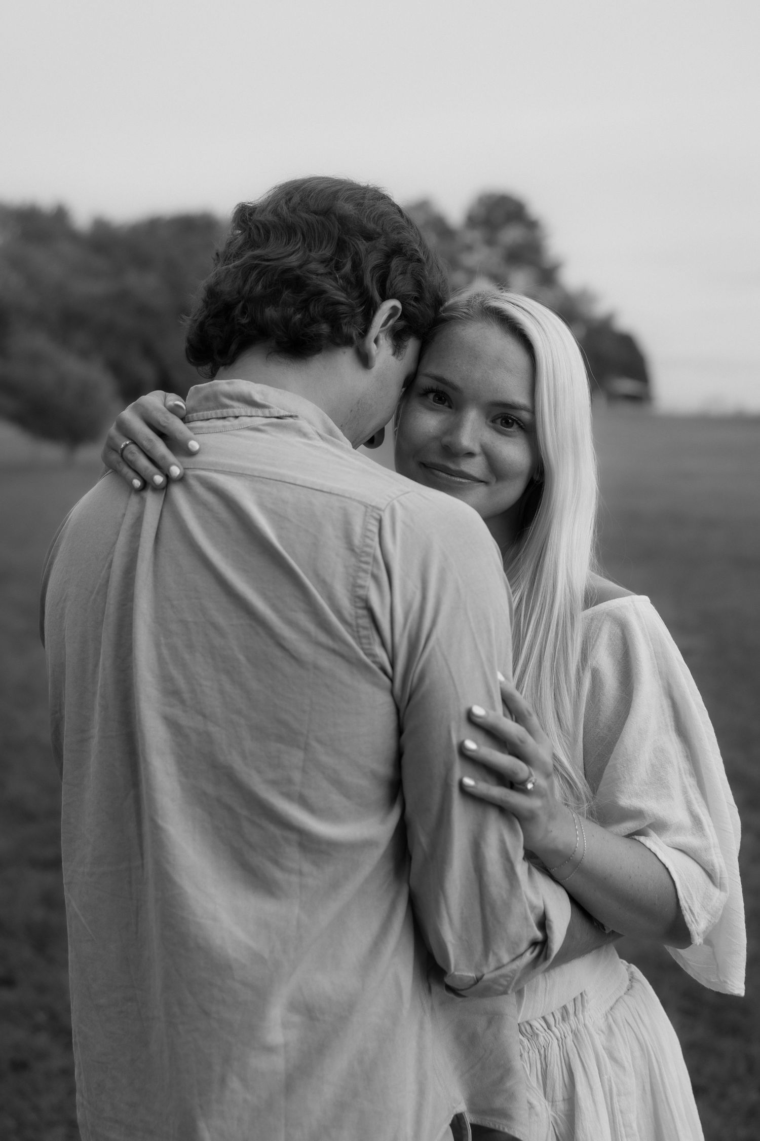 Black and white photo of a couple embracing in a field, woman smiling at camera while man kisses her cheek.