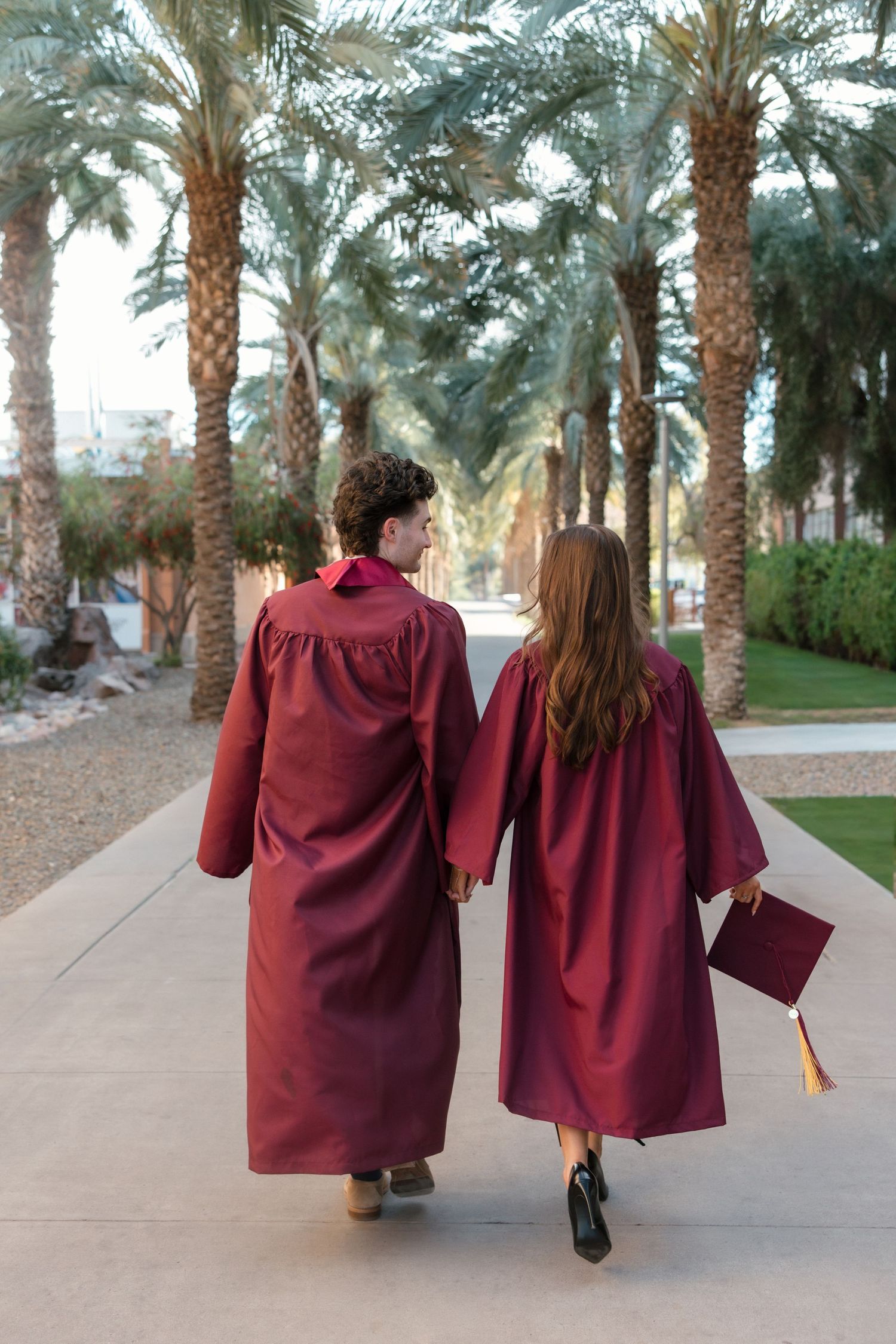 Two graduates in maroon caps and gowns walk hand in hand along a palm tree-lined path.