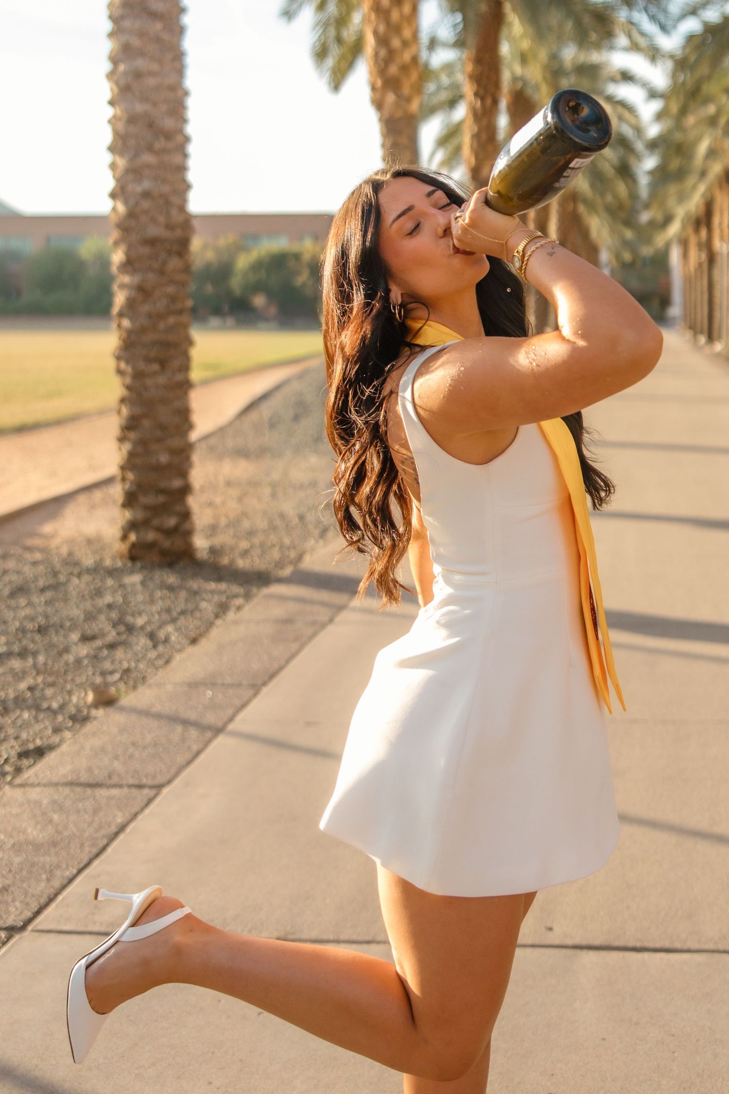 Young woman in white mini dress and heels drinking from champagne bottle outdoors near palm trees.