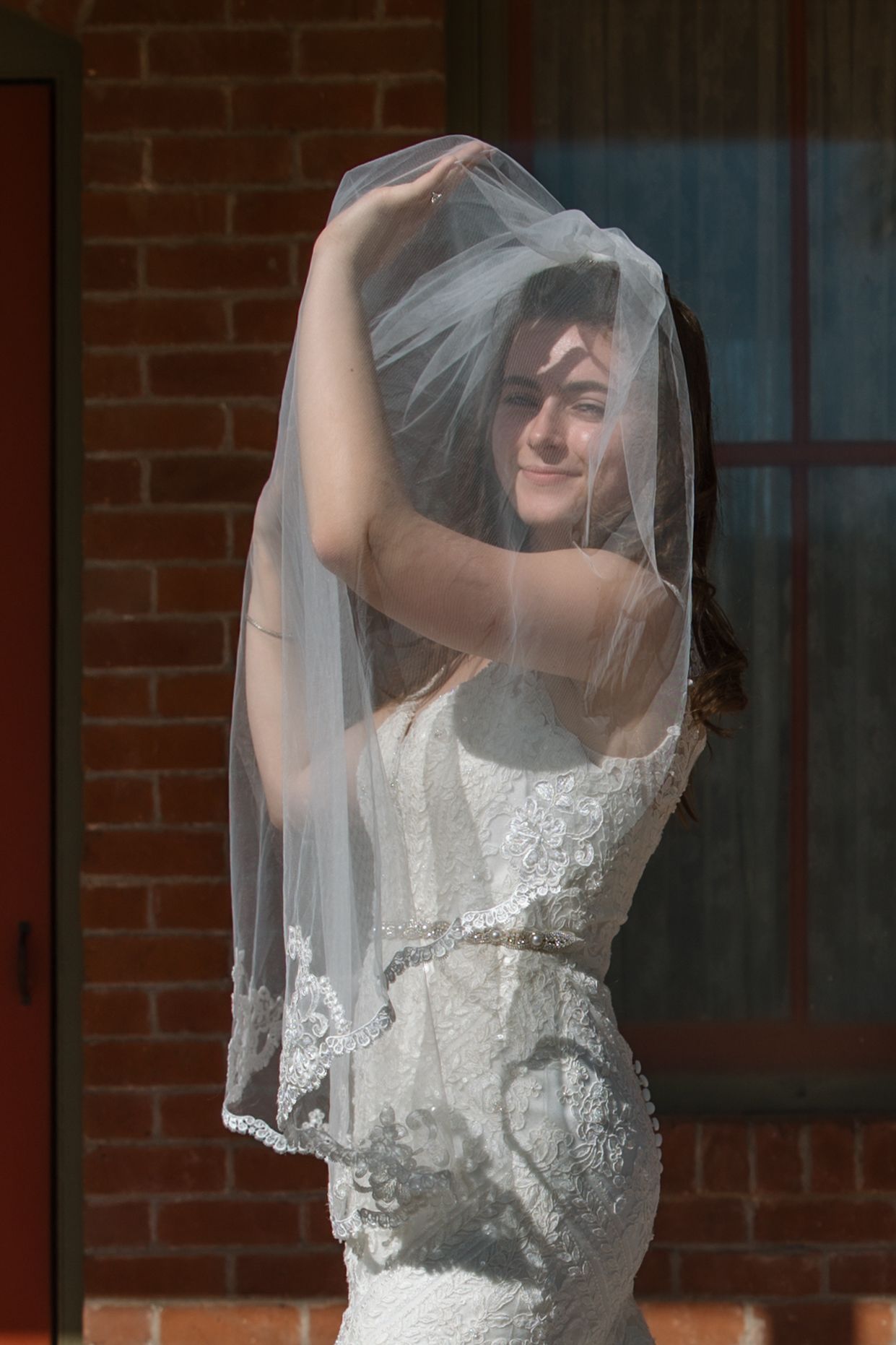 Bride lifts her lace veil over her head while smiling in front of brick wall.