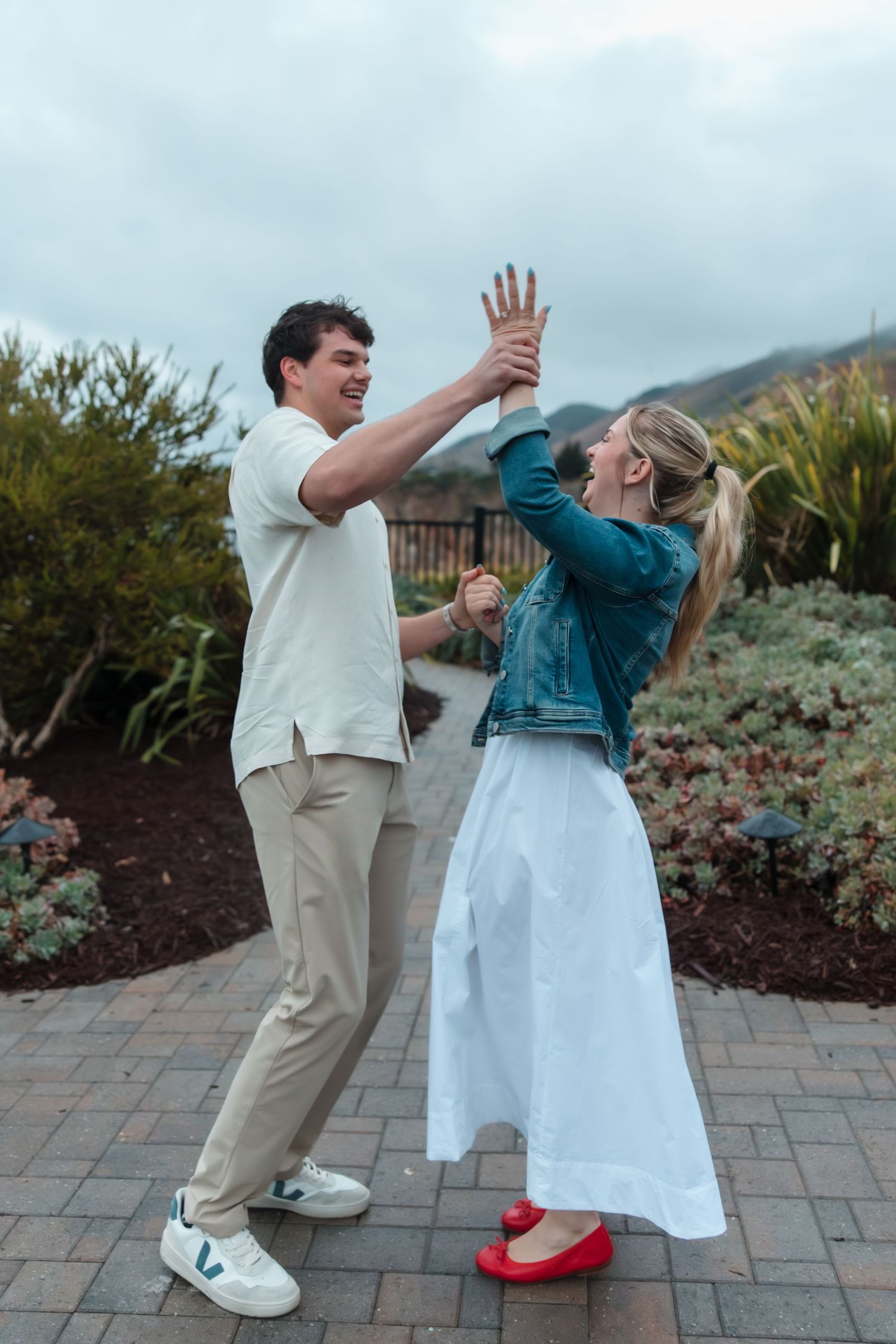 A joyful couple dancing outdoors on a brick path, woman in white skirt and red flats, man in white shirt and khakis.