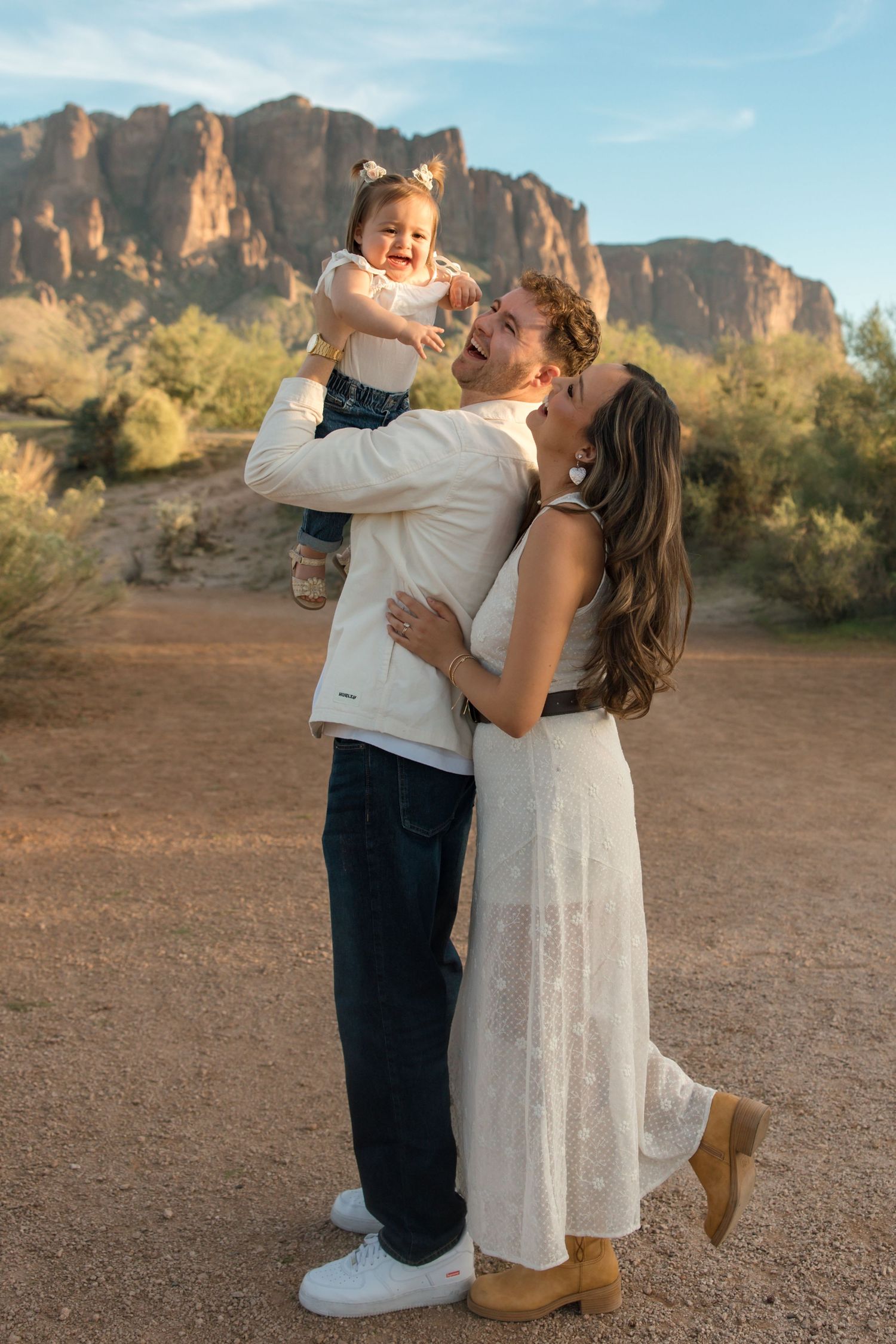 Joyful family moment with dad lifting laughing toddler as mom embraces them in the desert.