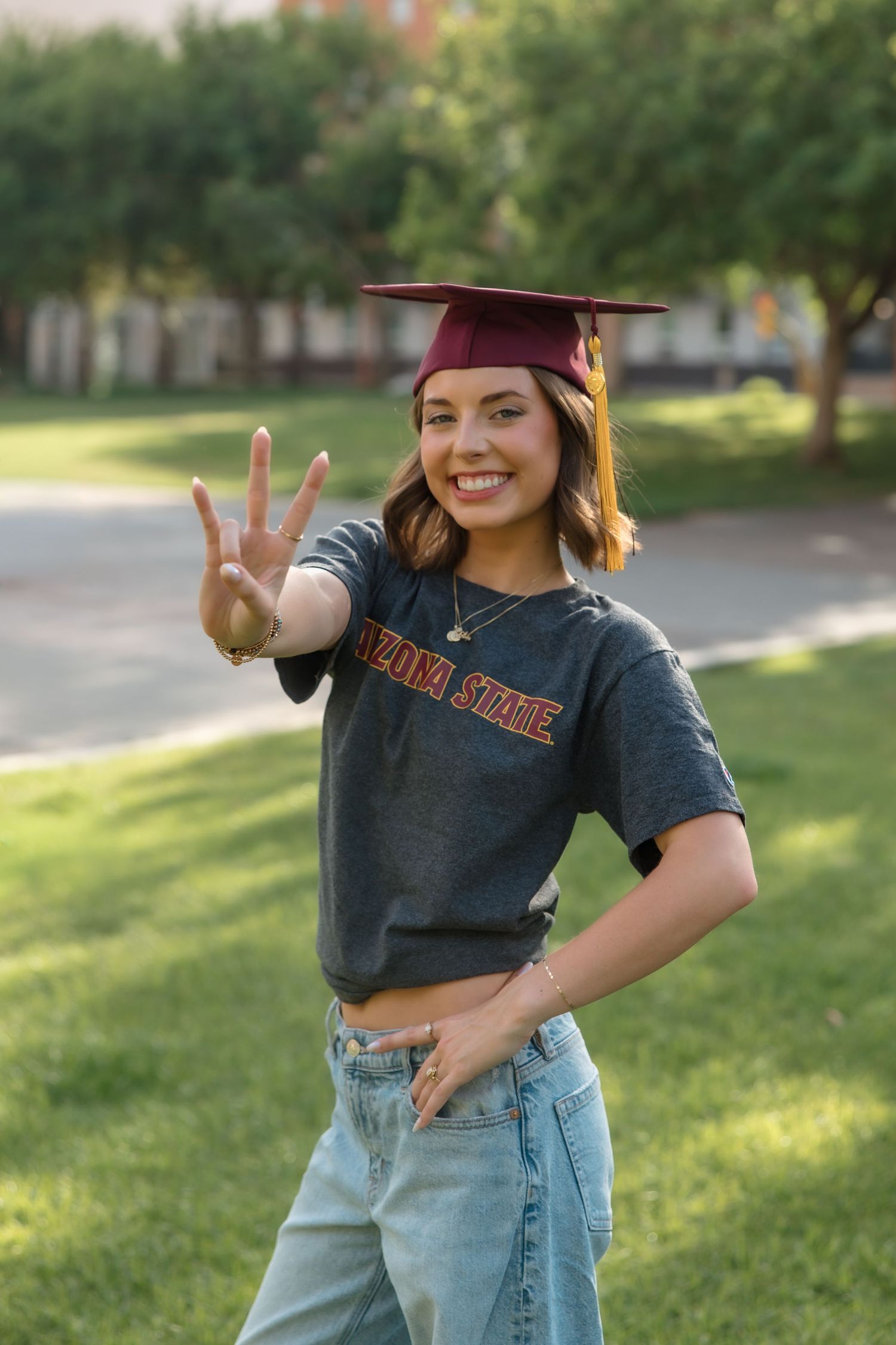 Smiling Arizona State University graduate in maroon cap and ASU t-shirt flashing pitchfork sign.