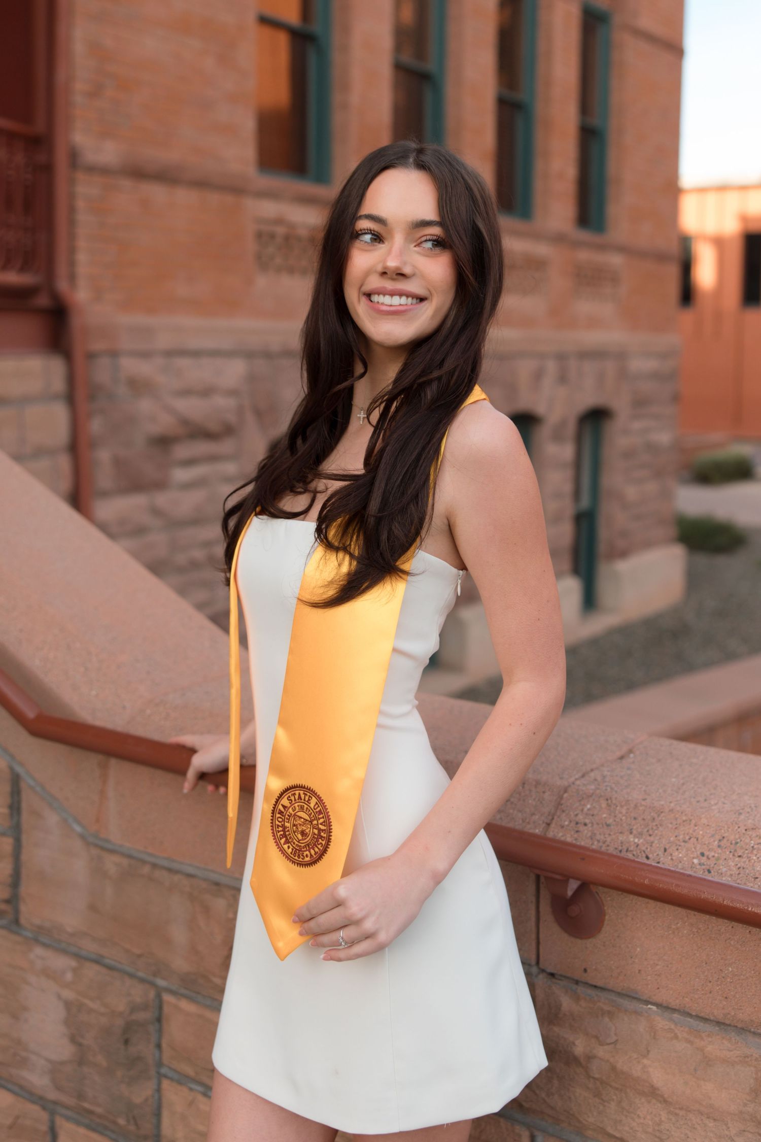 Brunette female graduate in white dress with gold graduation stole posing on brick campus balcony.