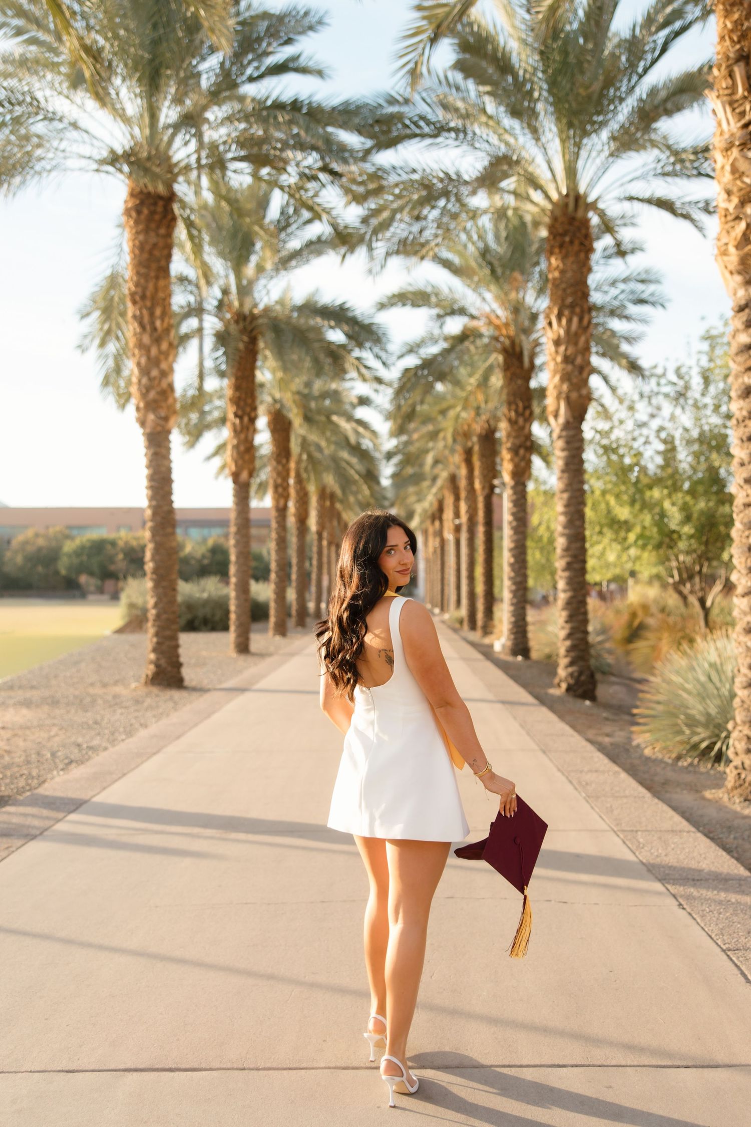 Female graduate in white backless dress walks along palm tree lined pathway holding maroon graduation cap.
