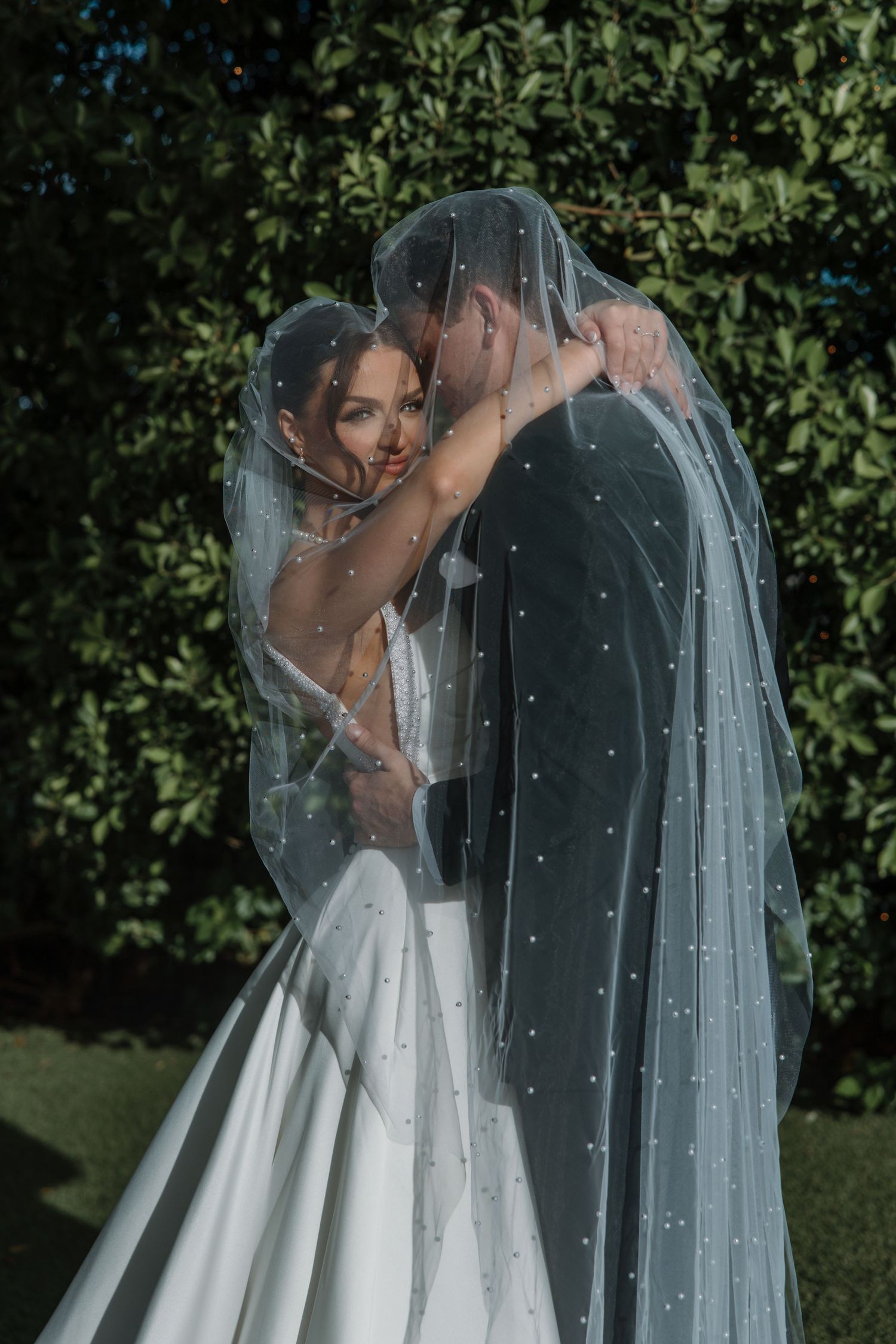 Bride and groom sharing a romantic moment under a pearl-embellished veil surrounded by green foliage.