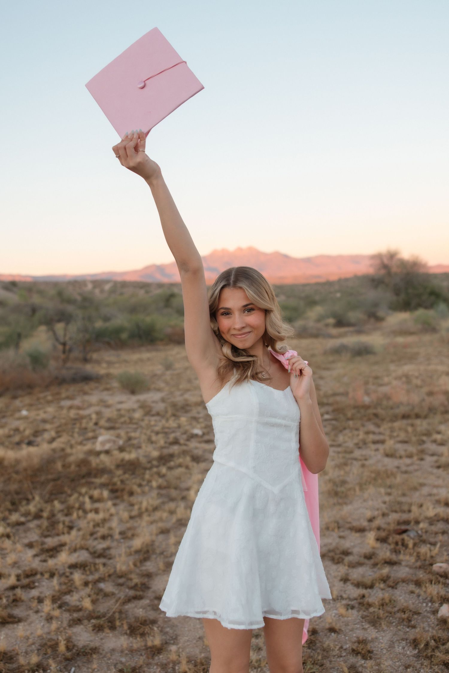 Young woman in white dress holding pink graduation cap aloft in desert landscape at sunset.