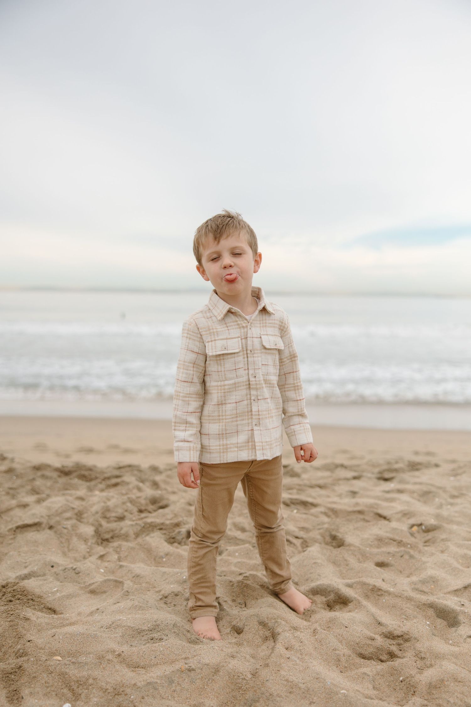 A young boy in a plaid shirt and tan pants stands barefoot on a sandy beach, sticking out his tongue.