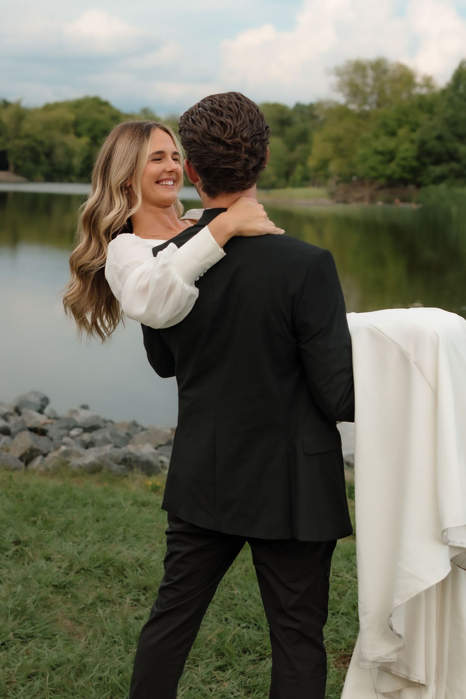 Joyful bride in white gown embraces groom in black suit near scenic lakeside with rocky shore.