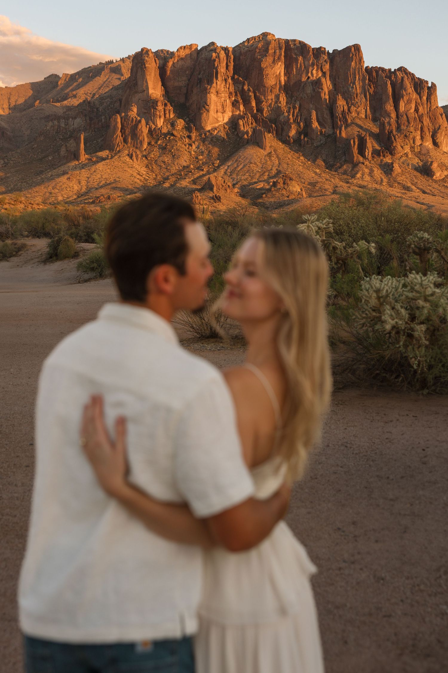 Couple embracing at sunset with Superstition Mountains glowing orange in the Arizona desert background.